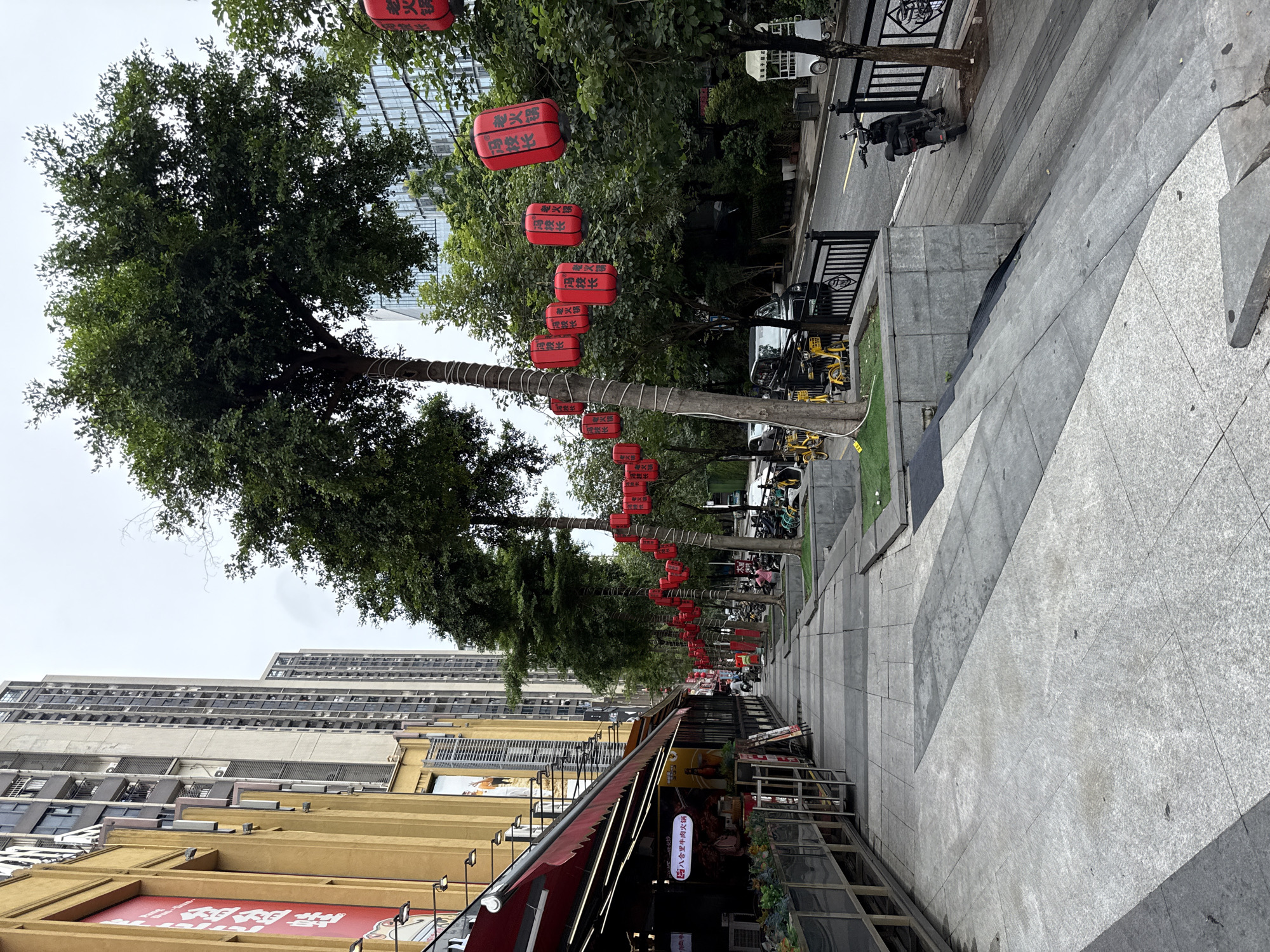The image depicts a street scene in Haizhu District, China, characterized by a wide, gray tiled sidewalk lined with trees decorated with red lanterns. To the left, a building with a yellow facade features awnings and signage, suggesting commercial establishments like restaurants or shops. The sidewalk leads past a row of trees, with several yellow rental bicycles parked to the right of the sidewalk. A street runs parallel to the sidewalk, with parked motorcycles and a vehicle visible. In the background, tall apartment buildings rise into a cloudy sky. The overall impression is a bustling urban environment with elements of both commerce and public amenity.