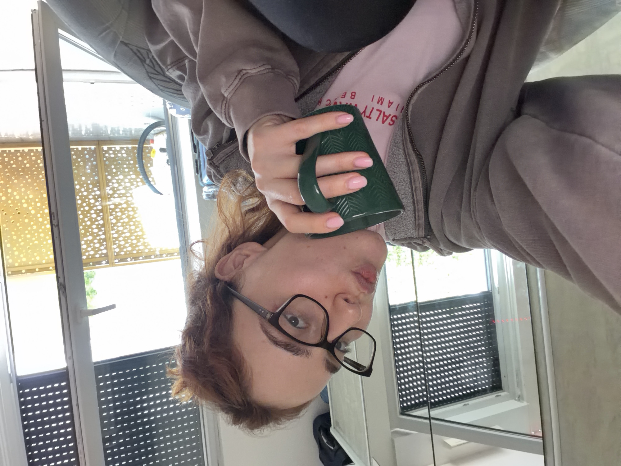 A young person with reddish-brown hair and glasses is holding a green mug with both hands. They are wearing a light pink t-shirt with red text and a grey zip-up hoodie. The person is looking towards the camera with a playful expression, their lips slightly puckered.
The scene appears to be indoors, near a window or door that leads to an outdoor area. Through the window, a bicycle is partially visible. The background shows a modern building with blinds or screens on the windows. The lighting suggests it is daytime. The overall mood is casual and relaxed.
Visible text on the t-shirt reads "SALTY MIAMI BE".