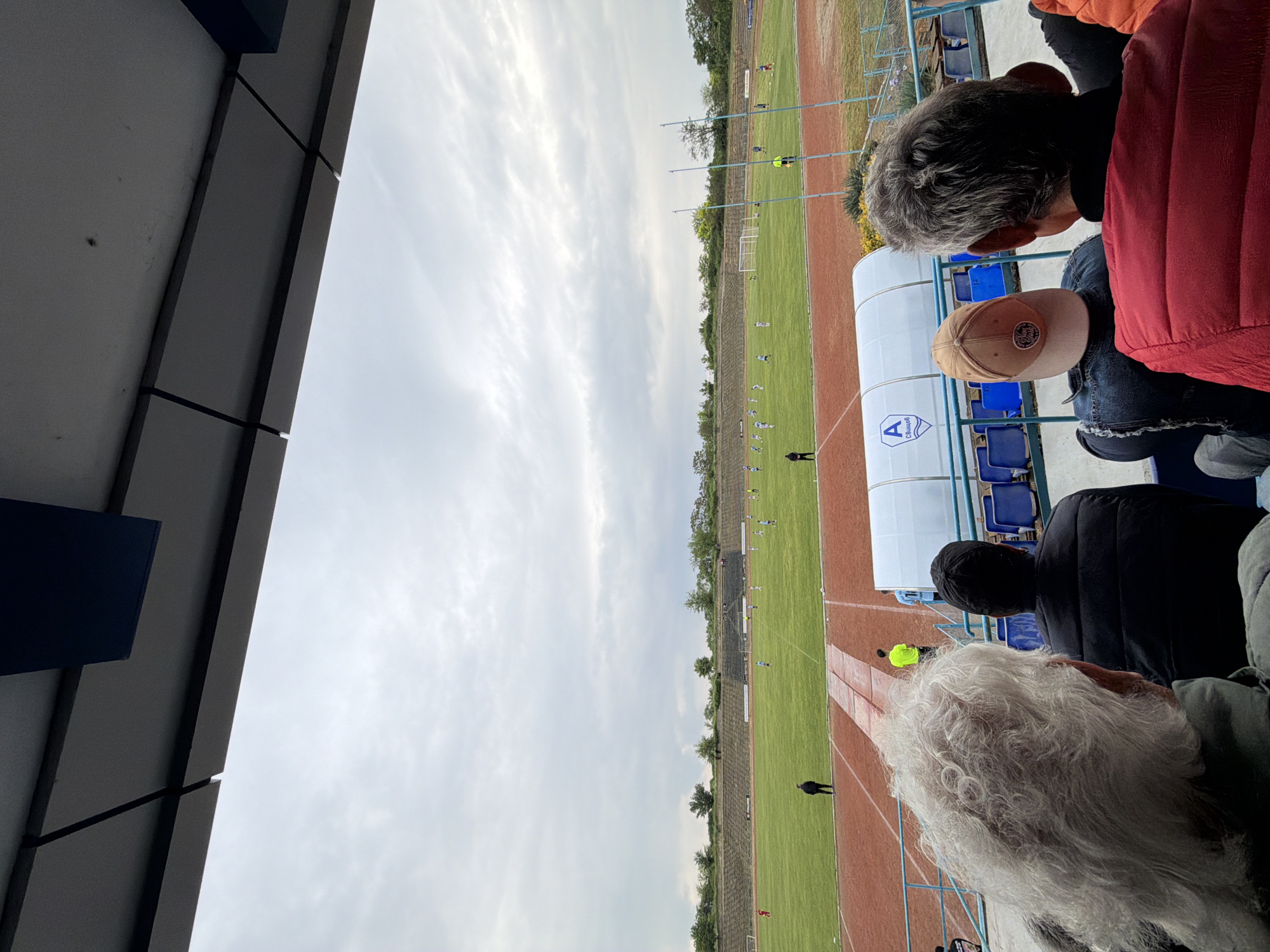 From a seated position within a stadium in Svishtov, Bulgaria, an observer views a soccer game in progress. Several people, including those with grey hair and a baseball cap, are seated in front. The field is lined with track lanes. A white structure with a logo is visible in the foreground beside the spectator seating. The sky is overcast.