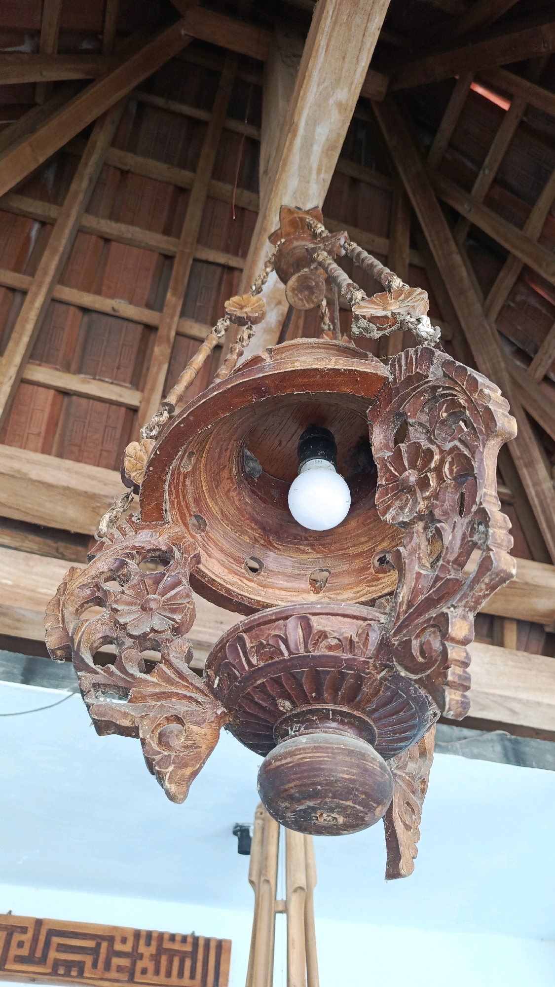 In Babadan, Indonesia, a vintage, intricately carved wooden lantern hangs from a wooden beam beneath a rustic, wooden-tiled roof structure. The lantern, made of dark wood, features elaborate floral carvings and a rounded, bell-like interior housing a white, modern LED light bulb. The wood appears weathered and aged, with some dust and cobwebs visible. The roof structure above consists of visible beams and reddish-brown tiles, suggesting an open-air or semi-open space. In the background, a white ceiling is visible, along with hints of bamboo structures and a wooden decorative element with Arabic calligraphy. The lighting is ambient, with the LED bulb being the primary light source from the lantern itself.