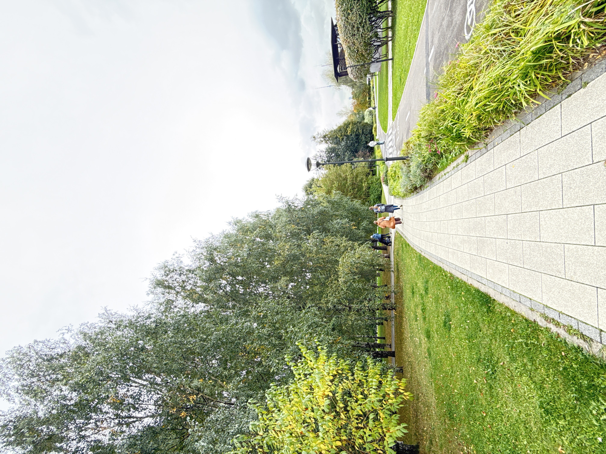 This is an image of a park in Moscow, Russia, featuring a paved pathway with a gray border, lined by green grass on one side and a row of tall, slender trees on the other. The trees have green foliage, with some hints of yellow and orange leaves, suggesting it may be autumn. The pathway curves to the right, leading into the distance where a road with bicycle markings is visible. Further in the background, there are more trees, a lamppost, and a modern building with a dark roof. Two women are walking on the pathway, one in a light brown coat and the other in a darker jacket and pants. A third person in a white jacket is walking on the road. The sky is overcast and light gray. The scene depicts a tranquil, everyday moment in an urban park.