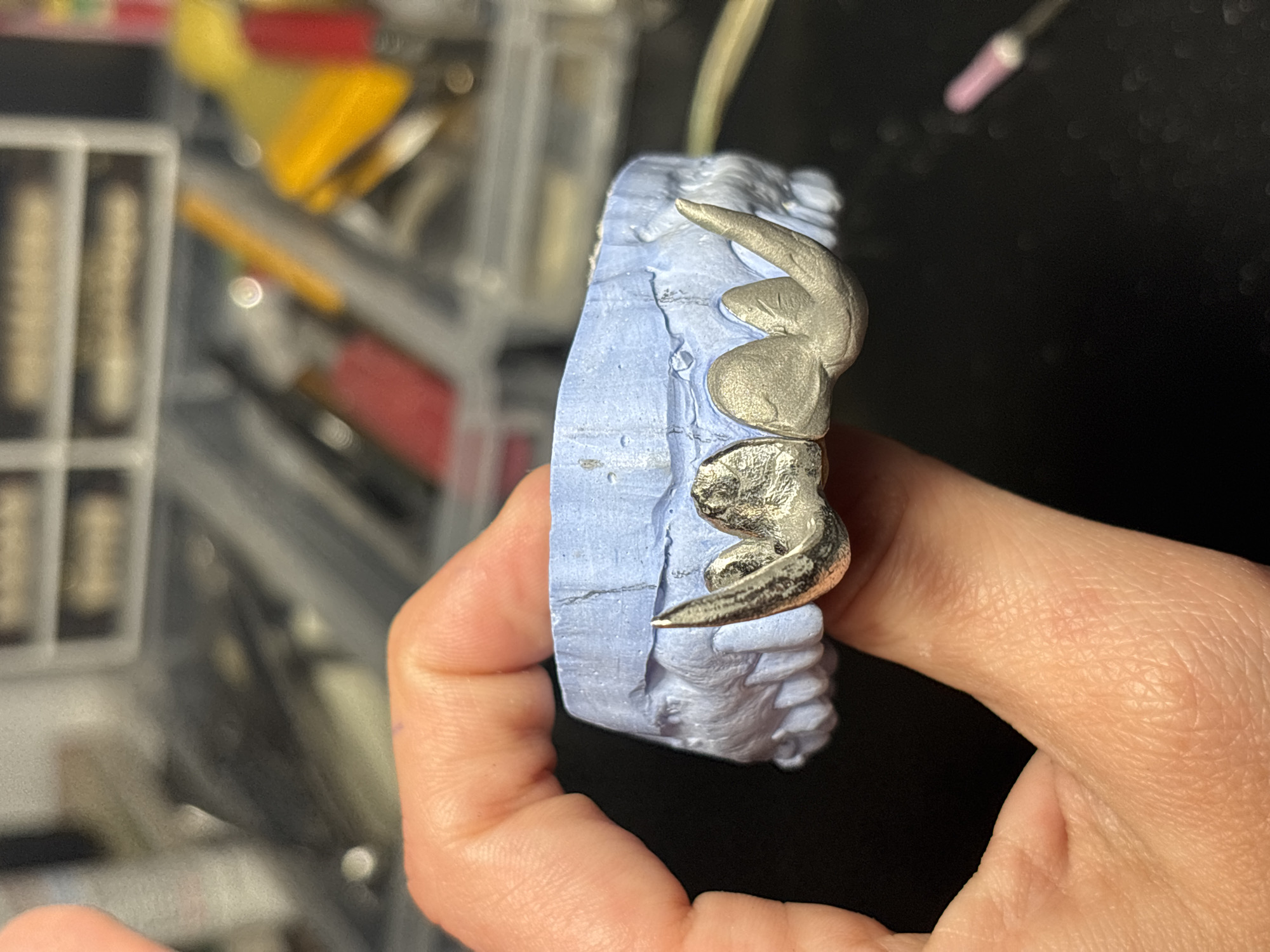 A hand is holding a dental mold with a metal dental restoration in place. The dental mold is light blue and captures an impression of teeth. The restoration is a silver-colored metal, likely a bridge or crowns with clasps. The background is blurry, but includes dental instruments and tools in a tray and on a black surface. There is nothing to suggest this image was taken in Sofia, Bulgaria.