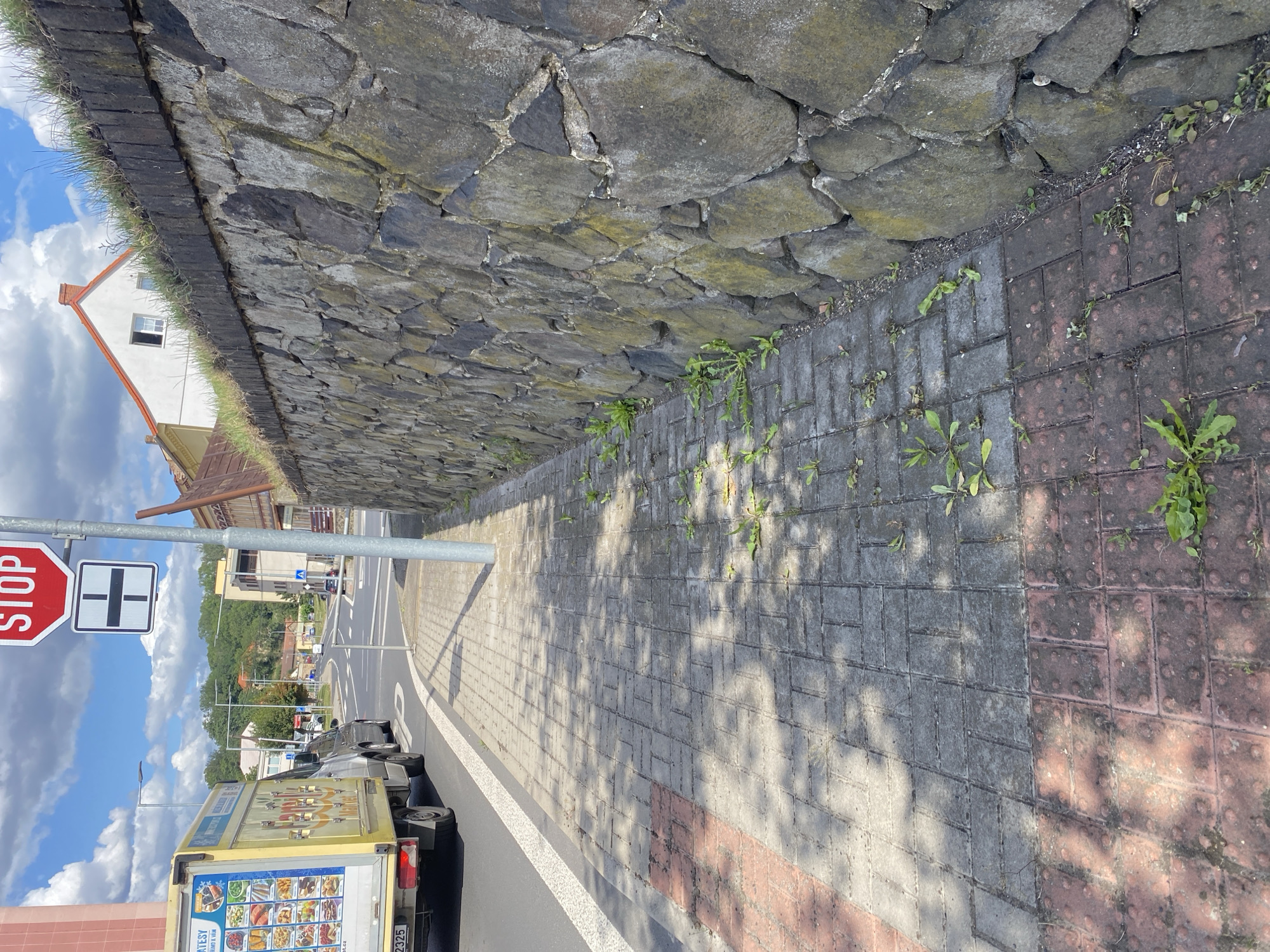 The image captures a street scene in Bílina, Czechia. A yellow delivery truck is parked on the left side of the road, its side displaying various food items. Adjacent to the road is a sidewalk paved with interlocking gray and red bricks, some of which are textured with raised dots. Weeds are growing through cracks in the sidewalk. A tall stone wall borders the sidewalk on the right, with a white house visible beyond it at the top of the wall. A street sign with a red octagon and the word "STOP" is mounted on a pole. Further down the street, other vehicles and buildings are visible. The sky is blue with scattered white clouds.