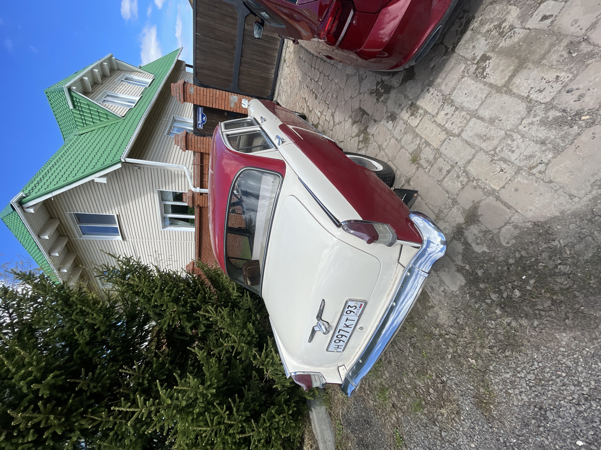 The image shows a two-tone, vintage car, predominantly white with a red roof, parked in what appears to be a driveway or paved area. The car's license plate is visible and reads "H997KT93 RUS". A portion of a red car is visible parked beside the vintage car. In the background, there's a two-story house with light-colored siding and a green roof. A dense evergreen bush is situated near the car, partially obscuring the view. The location appears to be Минино, Russia. A sign labeled "ул. Бурниковский" ("Burnikovskiy St.") is visible attached to the brick pillar next to the gate. The sky is blue with some clouds. The ground is paved with stone-like pavers.