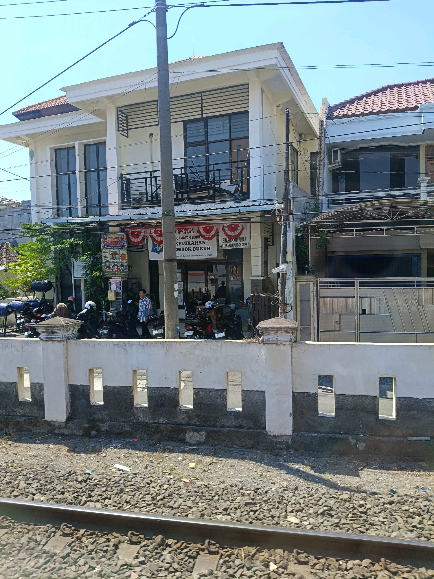The image depicts a street scene in front of a two-story building, likely in Surabaya, Indonesia, as suggested by text on a banner. The building is white with dark-trimmed windows and balconies. In front of the building, there is a low white wall with rectangular openings, behind which a collection of parked motorcycles is visible. A few individuals are present, some near the motorcycles and one standing on the sidewalk. Banners and signs adorn the building's facade, indicating a commercial or administrative establishment. The ground in front of the wall is covered in gravel and debris, with railway tracks in the foreground. The sky is clear and blue, suggesting a sunny day.