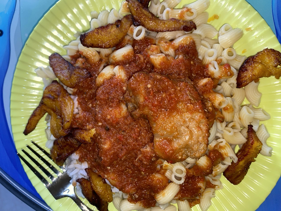 The scene depicts a prepared meal served on a bright yellow, ridged plastic plate. The dish consists primarily of white elbow macaroni pasta, largely covered by a thick, reddish-orange stew or sauce. Visible beneath portions of the sauce and next to the fork is a small amount of white rice. Two prominent pieces of protein, likely fish or chicken, are situated centrally, heavily coated in the red stew. Numerous pieces of golden-brown fried plantain are scattered across the pasta and around the perimeter of the plate. A silver-colored metal fork with four tines rests on the upper left rim of the plate. The yellow plate is positioned on a blue surface, possibly another tray or placemat, which features a subtle, wavy pattern. This meal is presented in a casual indoor setting in the city of Tema, Ghana.