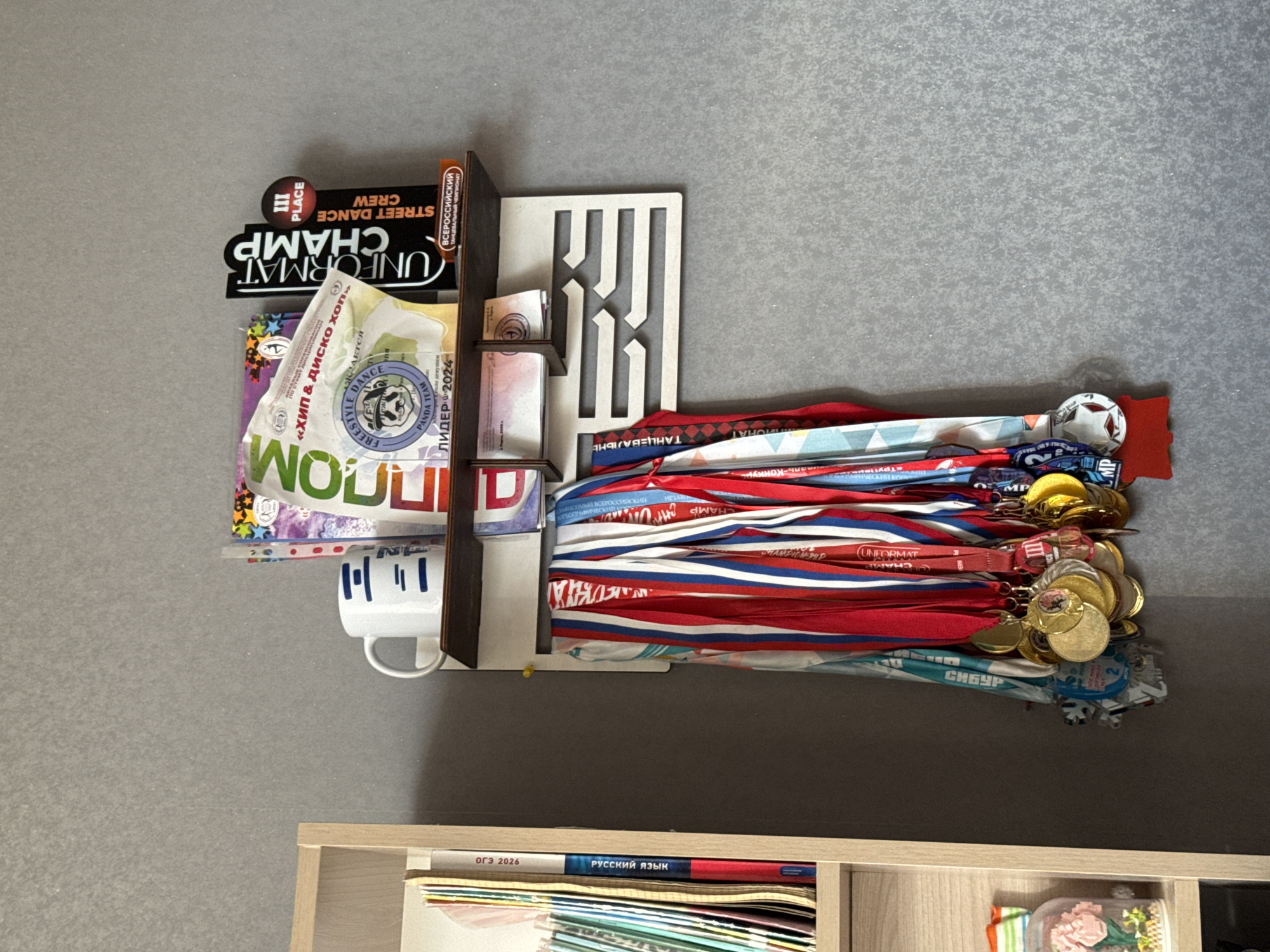 A wooden shelf is mounted on a gray textured wall, displaying several items. To the left, a light-colored wooden bookshelf is partially visible, containing several books. The shelf itself holds a white mug with blue lines, a stack of papers and documents, including a certificate or award with the text "HIP & DISCO HOP" and "Leader 2024", and a black sign that reads "UNFORMAT CHAMP STREET DANCE CREW" with a "III PLACE" emblem. Below the shelf, a collection of medals with various colored ribbons, predominantly red, white, and blue, hangs from a wooden rack. The medals appear to be awards for dance competitions. The overall scene suggests a display of achievements in dance. The location is not specified in the image.