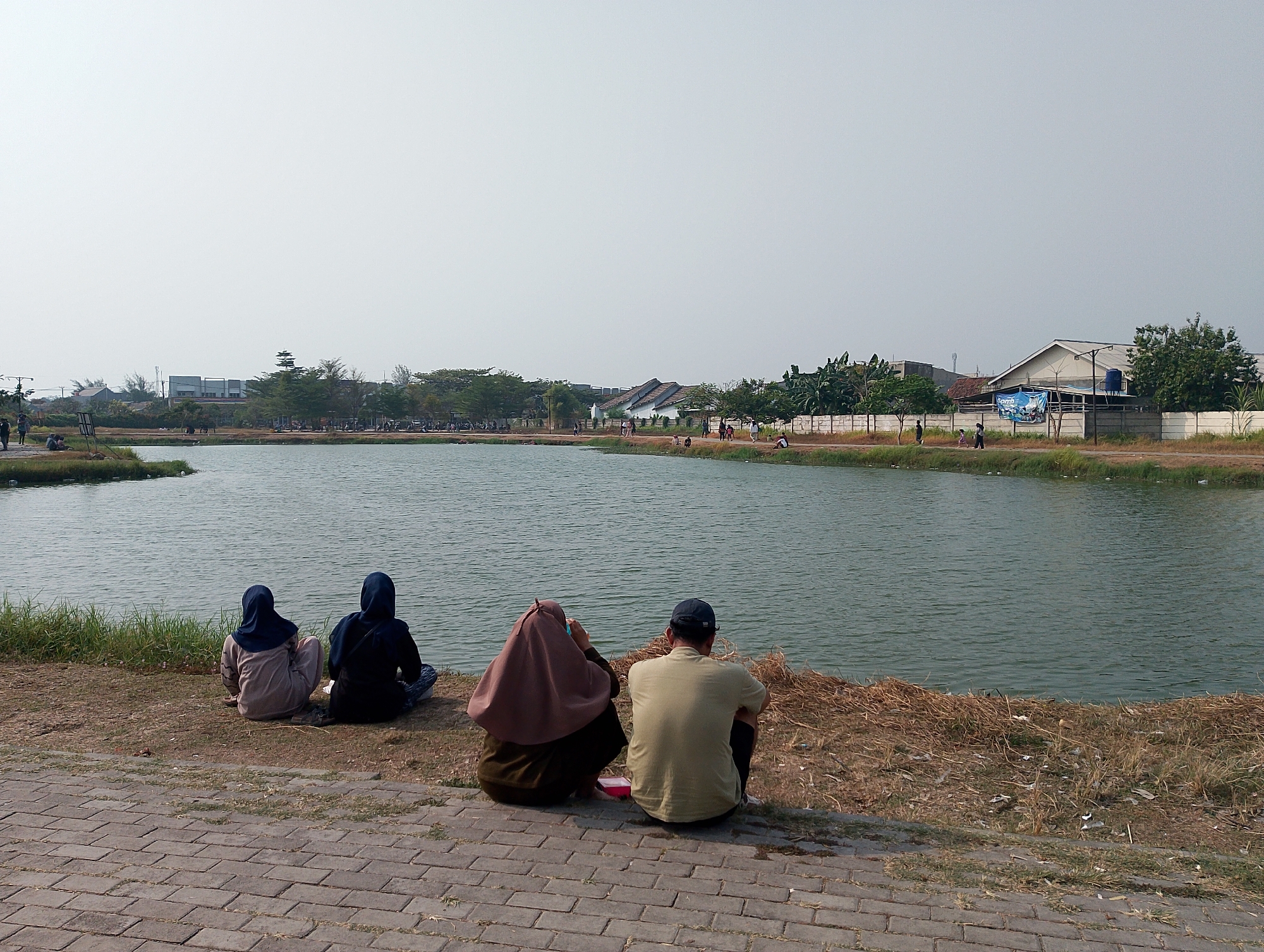 In the city of Karawang, Indonesia, a clear, bright day is depicted at a waterfront park. Four individuals are seated on the bank of a calm, green body of water, facing away from the camera and towards the lake. To the left, two women wearing hijabs are positioned close together. The woman closest to the camera wears a light brown hijab and a darker outfit. Next to her, a woman in a dark blue hijab and dark clothing is seated. To the right, a woman in a maroon hijab and dark clothing is seated, with a man wearing a light olive green t-shirt and a dark blue baseball cap next to her, also facing the water. A small red container is visible near the woman in the maroon hijab. The ground leading to the water is covered in dry grass and some debris, with a paved brick walkway in the foreground. Across the lake, buildings and trees are visible, suggesting an urban or semi-urban setting. The sky is overcast and a pale white color. The overall scene conveys a relaxed atmosphere of people enjoying a day outdoors by the water.
