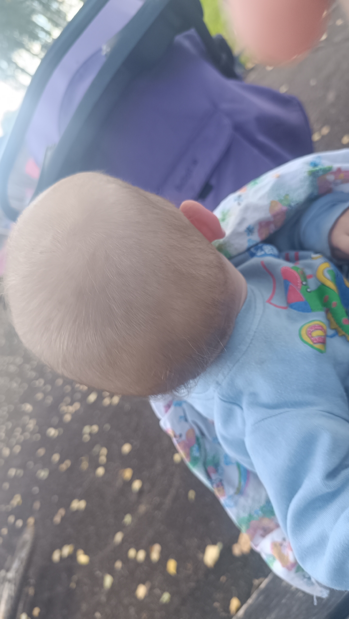 The image shows a close-up view of a baby's head and shoulders, with a portion of a stroller and blanket visible. The baby has light blonde hair and is wearing a light blue long-sleeved shirt with a colorful cartoon animal print visible on the blanket. The background is blurred, showing a dark surface with scattered yellow and brown spots that appear to be leaves or debris. The lighting suggests it is daytime, possibly outdoors. There is no indication of the location being Karsha, Russia.