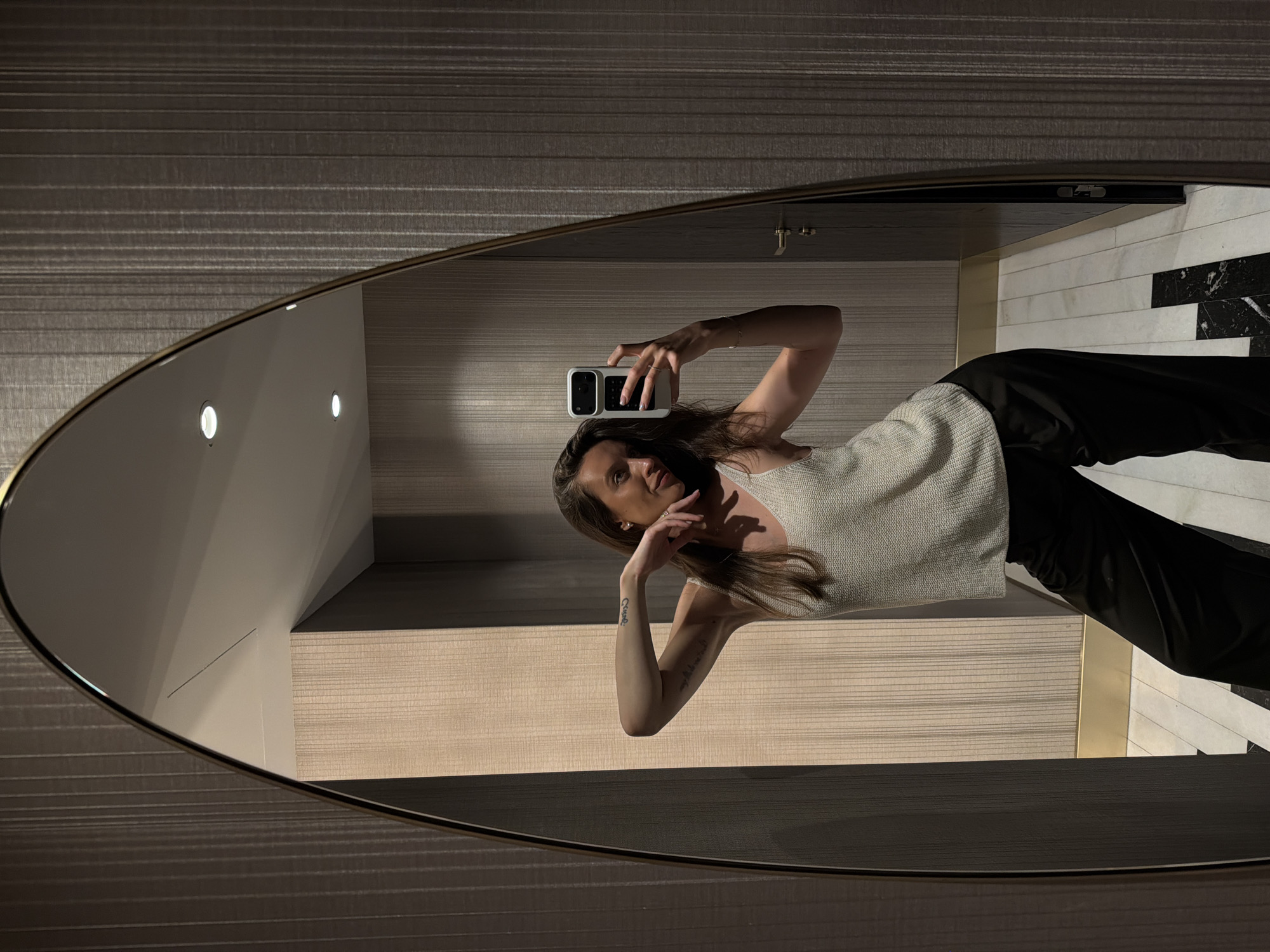 An adult female is captured in a mirror selfie within an indoor setting in Budva, Montenegro. The subject, positioned centrally in the mirror's reflection, has long, dark brown hair and is wearing a light-colored, textured sleeveless top and dark, wide-leg trousers. A tattoo with visible text is present on her right forearm. She is holding a white smartphone with a dark camera module in her right hand, pointed towards the mirror to capture the image. Her left arm is bent, with her hand near her chin. The mirror itself is irregularly shaped, elongated, and features a thin, dark frame.
The environment, visible both directly and in the mirror's reflection, consists of walls adorned with vertically striped textured wallpaper in varying shades of brown and beige. Recessed lighting fixtures are visible on the light-colored ceiling within the reflection. A dark-colored door with a metallic handle and lock mechanism is present on the right side of the reflection. The floor features a pattern of alternating white and black rectangular tiles, separated by thin grout lines. A gold-colored trim is visible at the base of the wall sections.