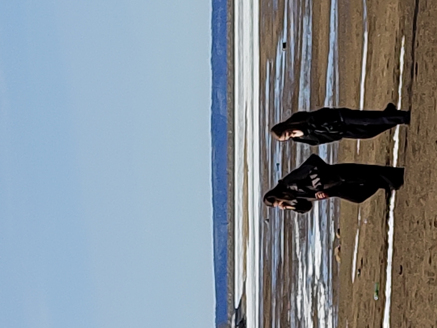 Two individuals clad in black clothing are positioned on a beach or shore, adjacent to what appears to be a body of water, possibly the Kama River. The location is most likely near the city of Chaikovsky, Russia, based on the landscape and potential access to the river. The individuals are standing or posing on the beach. The environment consists of a shore area possibly comprising sand or sediment, with a body of water in the background that appears to have a partially frozen surface. The sky is pale blue.