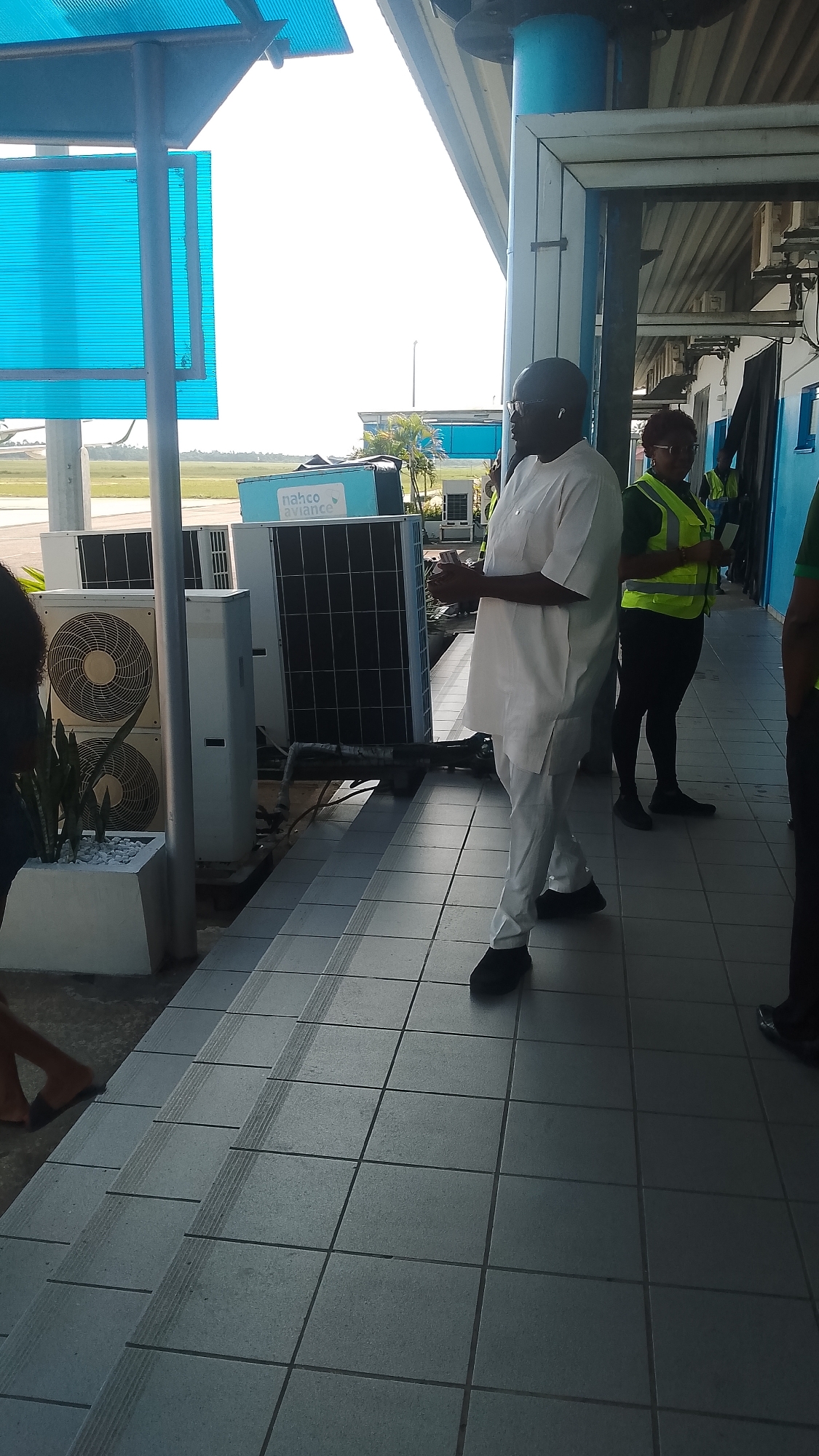 The image captures an outdoor scene at an airport in Orerokpe, Nigeria. In the foreground, a tiled walkway leads to a set of stairs. Several large air conditioning units are visible, some stacked vertically. A light blue container with the "nahco aviance" logo is also present. To the right, a man wearing a white traditional outfit and glasses stands looking to his left. Behind him, a woman in a neon yellow vest is visible. The background shows an airfield with palm trees and buildings under a bright, overcast sky. The overall impression is one of an active airport environment.
