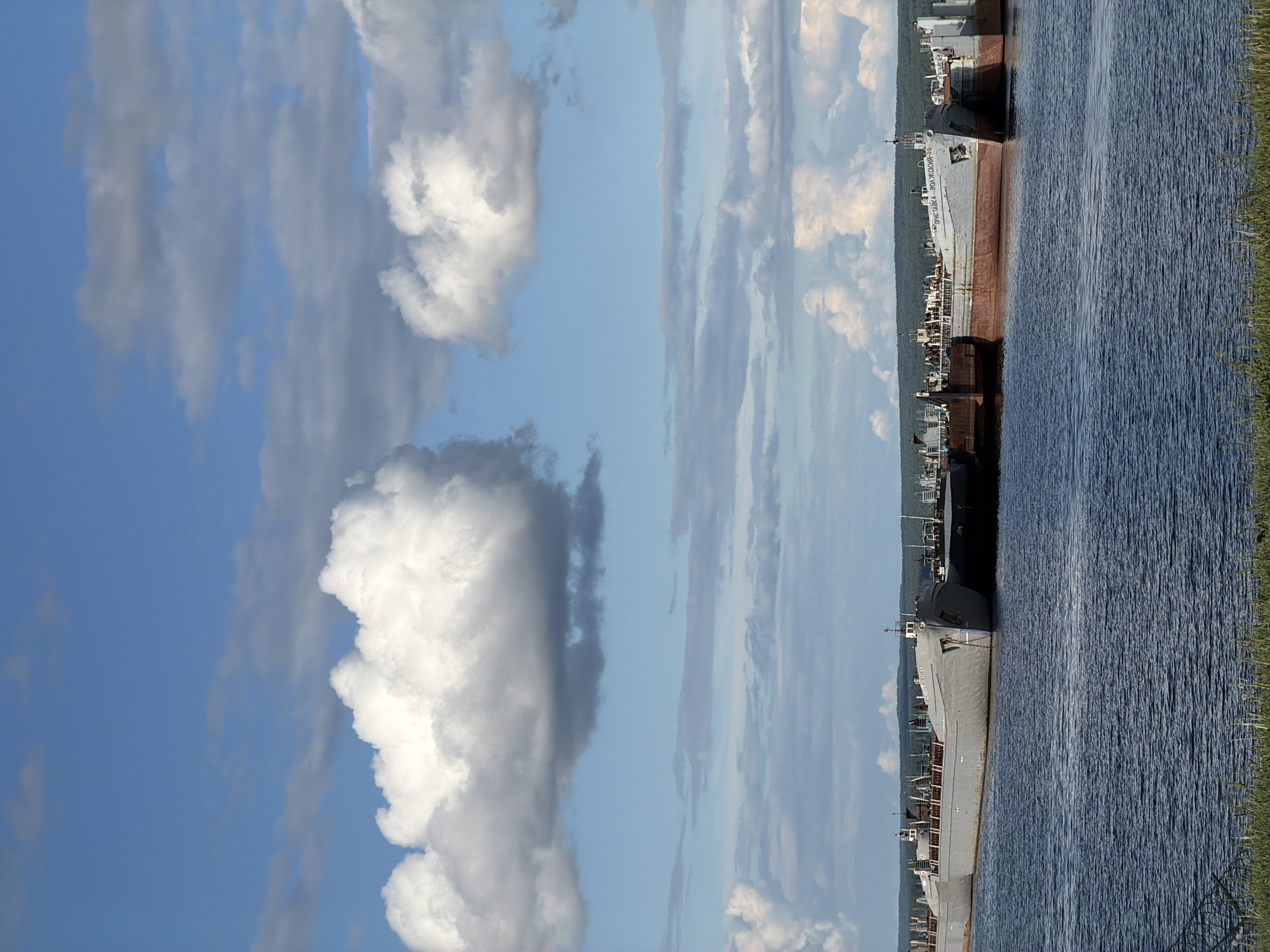 The image depicts a scene near the city of Chaikovsky, Russia, where a row of ships are docked along a waterway. The water, with visible ripples, is bordered by green vegetation on the far right. To the left of the waterway, several ships are positioned side by side. In the background, there are buildings and trees indicating the presence of the city. The left portion of the image features a bright blue sky with scattered white and grey clouds.