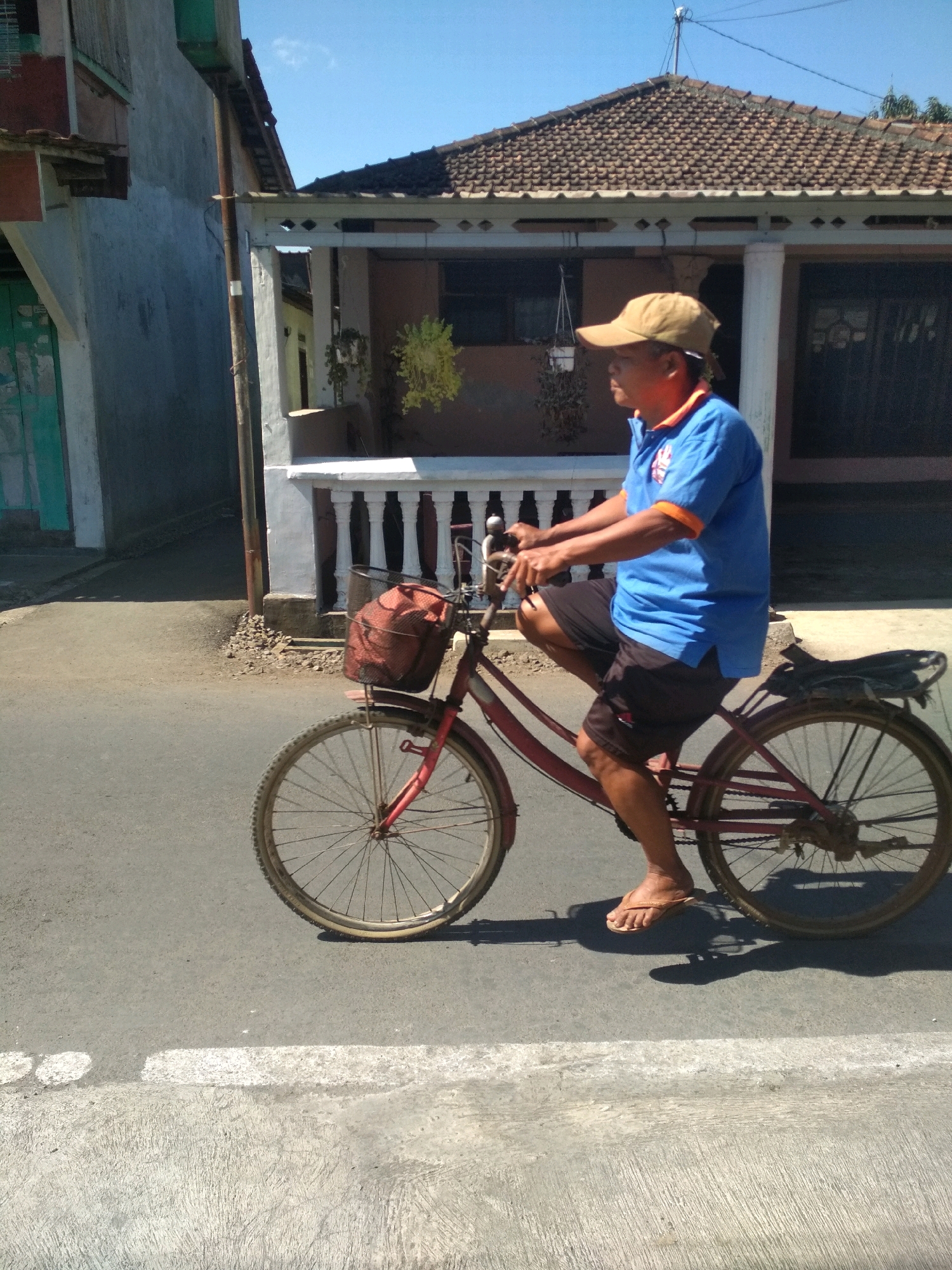 A man wearing a blue polo shirt and a tan baseball cap rides a red bicycle on a paved road in Desa Pamijen, Kecamatan Sokaraja, Indonesia. The man is in profile, facing left. He has dark skin and is wearing dark shorts and flip-flops. His left leg is extended forward, pedaling. A black wire basket with a red bag is mounted on the front of the bicycle. Behind the man, a house with a tiled roof and white railings is visible. The house has a brown exterior and a dark-colored door. The sky is clear and blue. The road has a white line painted on the right side of the frame. The lighting suggests it is daytime.