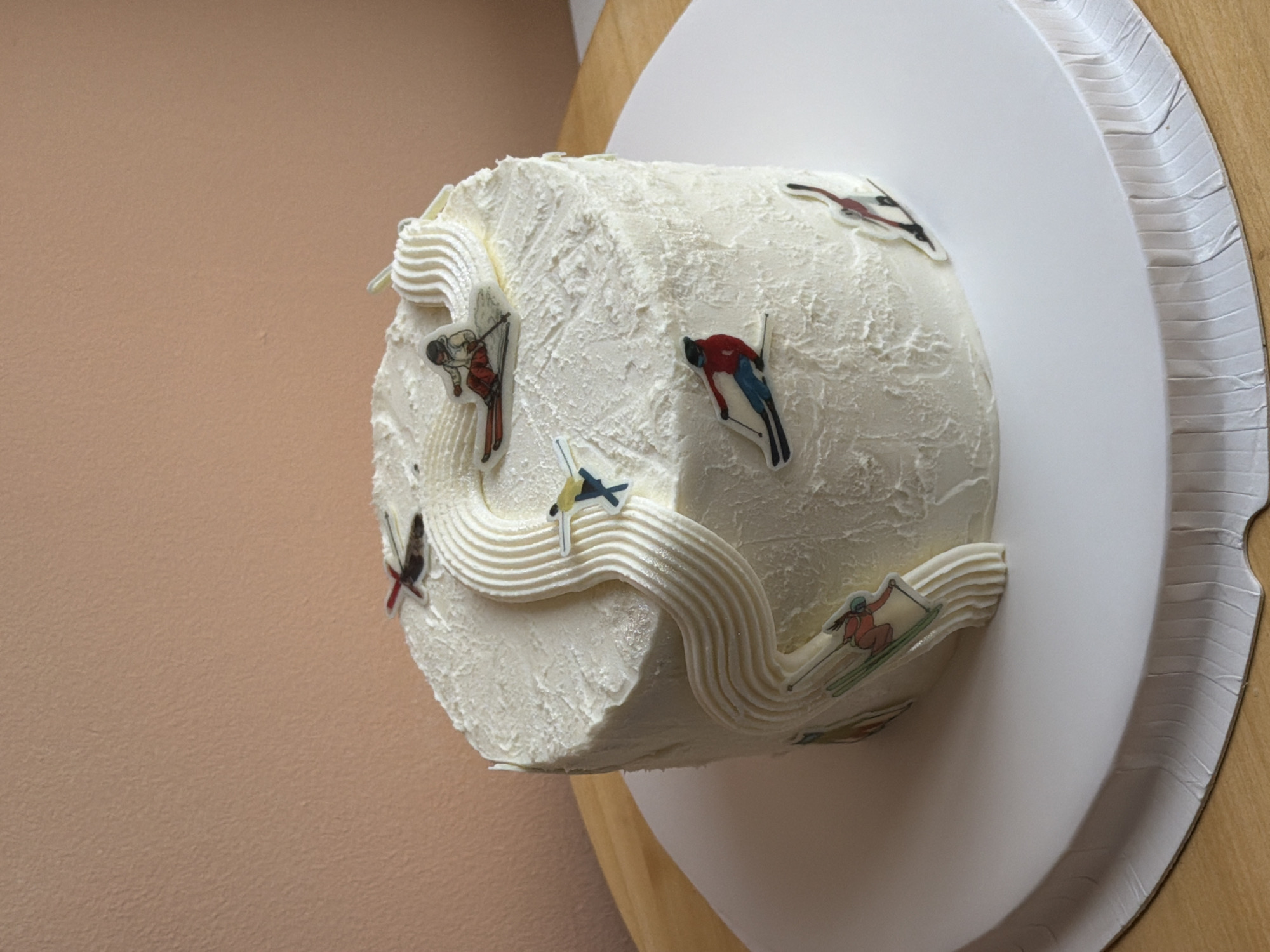 This media file presents a round cake, creatively decorated with a skiing theme. The cake is entirely covered in thick white frosting, which has been textured to resemble snow or ice, giving it a rugged, wintry appearance. Wavy lines of piped white frosting undulate across the cake's surface, mimicking ski slopes or snowdrifts.
Strategically placed on and around these frosting features are several small, colorful cutout figures of skiers in various dynamic poses, some appearing to ski down the frosting tracks, others scattered across the snowy texture. The figures depict skiers in different colored outfits, some with ski poles and skis, adding a sense of movement and sport to the dessert.
The cake rests on a white, circular cake board, which has a subtly ridged or crimped edge. This board is positioned on a light wooden surface, partially visible on the right side of the frame. A plain, light beige or tan-colored wall serves as the background to the left of the cake. The scene is illuminated by what appears to be bright indoor lighting. There is no visible text in the image. This photograph was taken in Tel-Aviv, Israel.