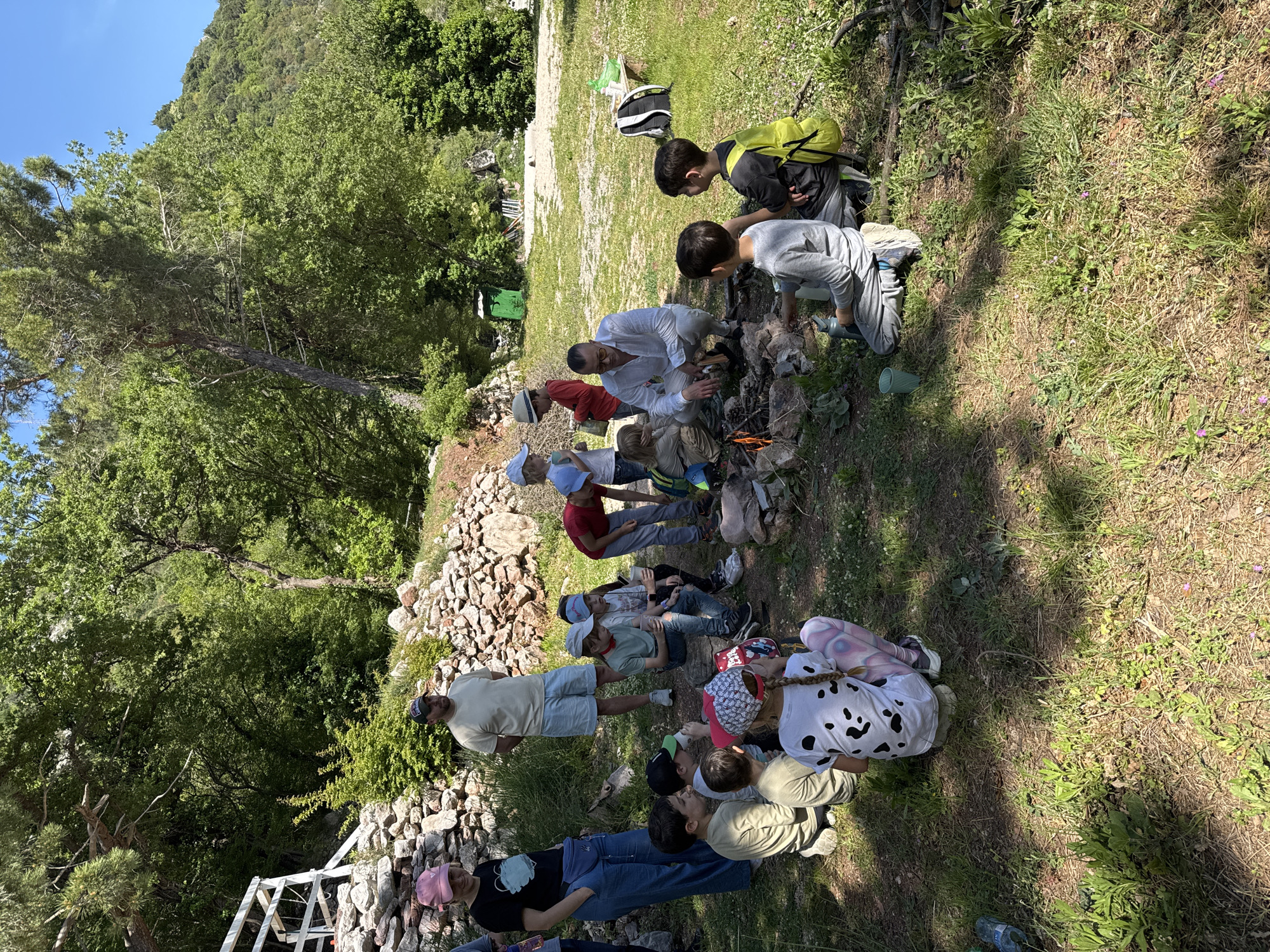 A group of approximately fifteen people, comprising adults and children, are gathered outdoors in a sunny, natural setting. The main activity centers around a small campfire burning within a ring of stones, located in the mid-ground. An adult male wearing a white shirt and glasses is actively tending to the fire, while a child in a grey long-sleeved shirt holds a green water bottle nearby, possibly pouring its contents. Several other children, including two with colorful backpacks (one yellow and green, one black), stand or sit close to the fire, observing with interest.
On the left side of the scene, more children and at least one adult are seated or standing on the grassy ground. One child in the foreground wears a distinctive white top with black spot patterns. An adult male in a light t-shirt and shorts stands among them. The adults and children appear engaged and relaxed, suggesting a recreational or educational outdoor activity.
The setting is characterized by lush green foliage, with numerous trees and hills visible in the background under a clear, bright blue sky. The ground is an uneven mix of grass, dirt, and scattered stones, including a low dry-stone wall that runs along the left side of the gathering. Strong sunlight indicates it is likely midday or early afternoon. No discernible text is visible within the image.