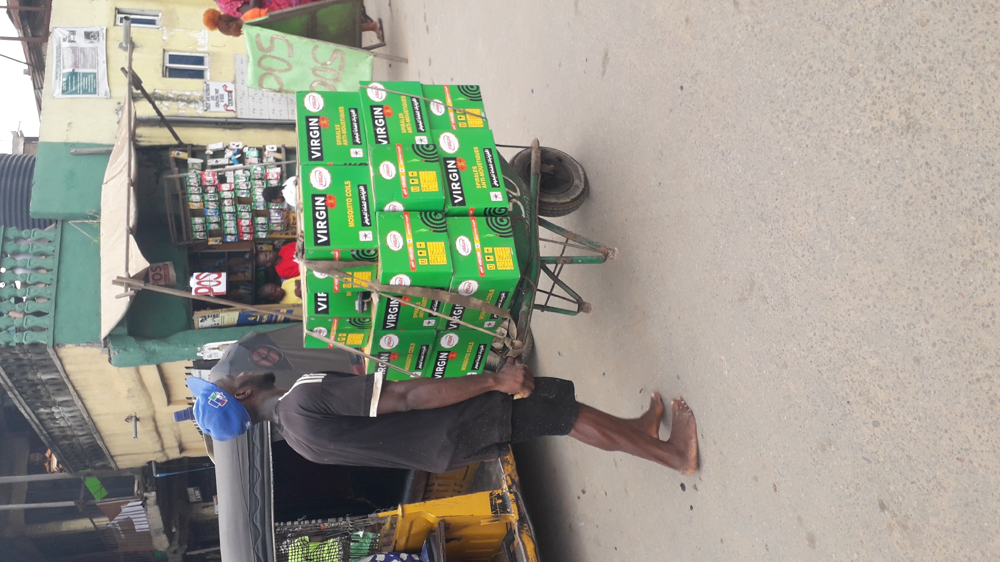 A man is pulling a wheelbarrow filled with boxes of "Virgin Mosquito Coils" down a city street. He is wearing a blue baseball cap, black shirt with white stripes on the sleeves, and black shorts. He is barefoot. The wheelbarrow is a faded green color. Behind the man, there is a yellow auto rickshaw. In the background, there are buildings and shops. "POS" is visible in the background. This image appears to be taken in a commercial area of Onitsha, Nigeria.
