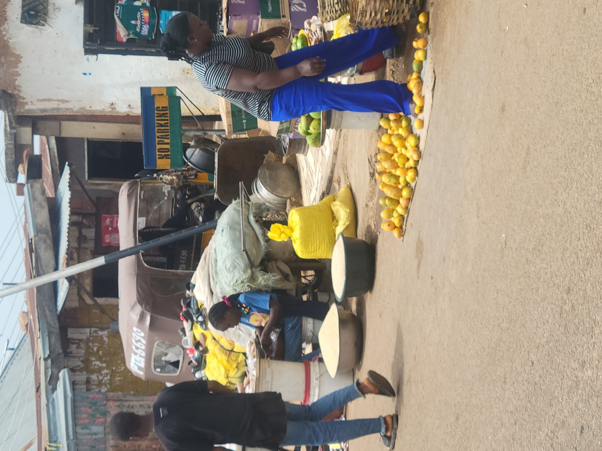 The image captures a vibrant street scene in Jos, Nigeria, showcasing everyday life around a roadside market. An African woman wearing a striped shirt and blue pants is positioned behind a display of fresh mangoes, possibly a vendor. A younger girl in a blue shirt appears to be involved in the market activity, perhaps assisting with tasks like sorting or preparing items. Another person wearing a black shirt and jeans is walking nearby, interacting with the environment.
Various objects are present, including buckets, bags of goods (one notably yellow), and containers used for displaying or storing market items. A "NO PARKING" sign is visible in the background, along with a tricycle (auto-rickshaw) with the license plate "PUA-6690," suggesting this is a bustling, active street. The scene has a cluttered, informal arrangement characteristic of street markets in many urban areas, with items spilling onto the pavement and creating a busy atmosphere. The environment conveys an everyday moment in a vibrant Nigerian city.