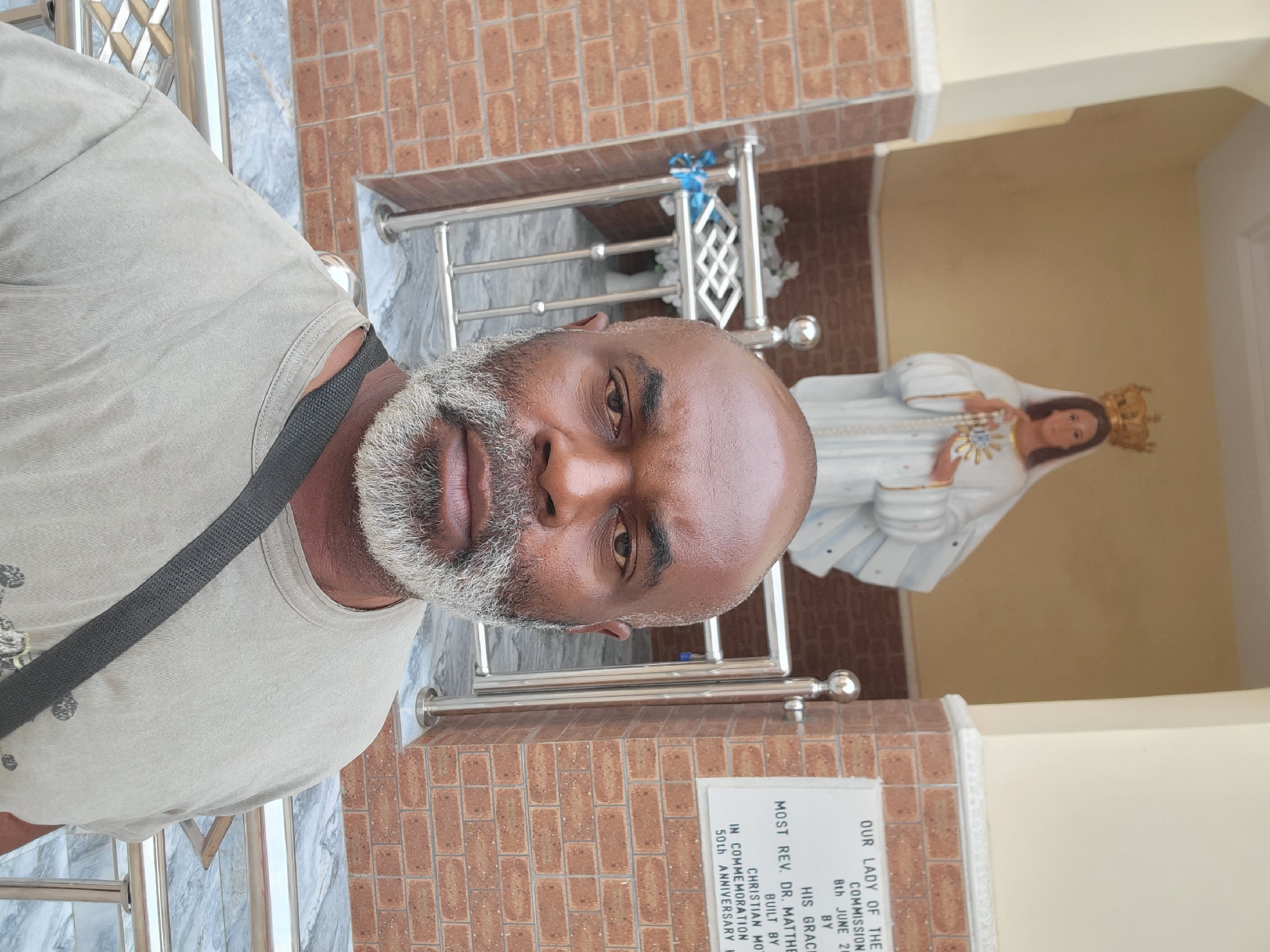A vertical photograph taken in Maiburiji, Nigeria, features a man in the foreground alongside a statue of the Virgin Mary in the background. The man, positioned on the left side of the frame, has dark skin, a bald head, and a full, prominent gray beard and mustache. He is wearing a light-colored t-shirt with a dark strap across his shoulder and gazes directly at the camera with a neutral expression.
Behind him, to the right, is a statue of the Virgin Mary, depicted in a white robe with gold trim, a golden crown, and holding a heart with a halo on her chest. The setting appears to be a commemorative or religious space, characterized by a brown brick wall in the upper background, grey marble-patterned flooring, and shiny silver railings. The bright, natural lighting suggests it is daytime.
A white plaque below the statue is partially visible, containing text that reads: "OUR LADY OF THE COMMISSION 8th JUNE 20 BY HIS GRACI MOST REV. DR. MATTHE BUILT BY CHRISTIAN MO IN COMMEMORATION 50th ANNIVERSARY". This inscription indicates the statue was commissioned or dedicated on June 8th, likely by Most Rev. Dr. Matthew and built by Christian Mo, to commemorate a 50th anniversary. The overall scene conveys a sense of calm and reverence.