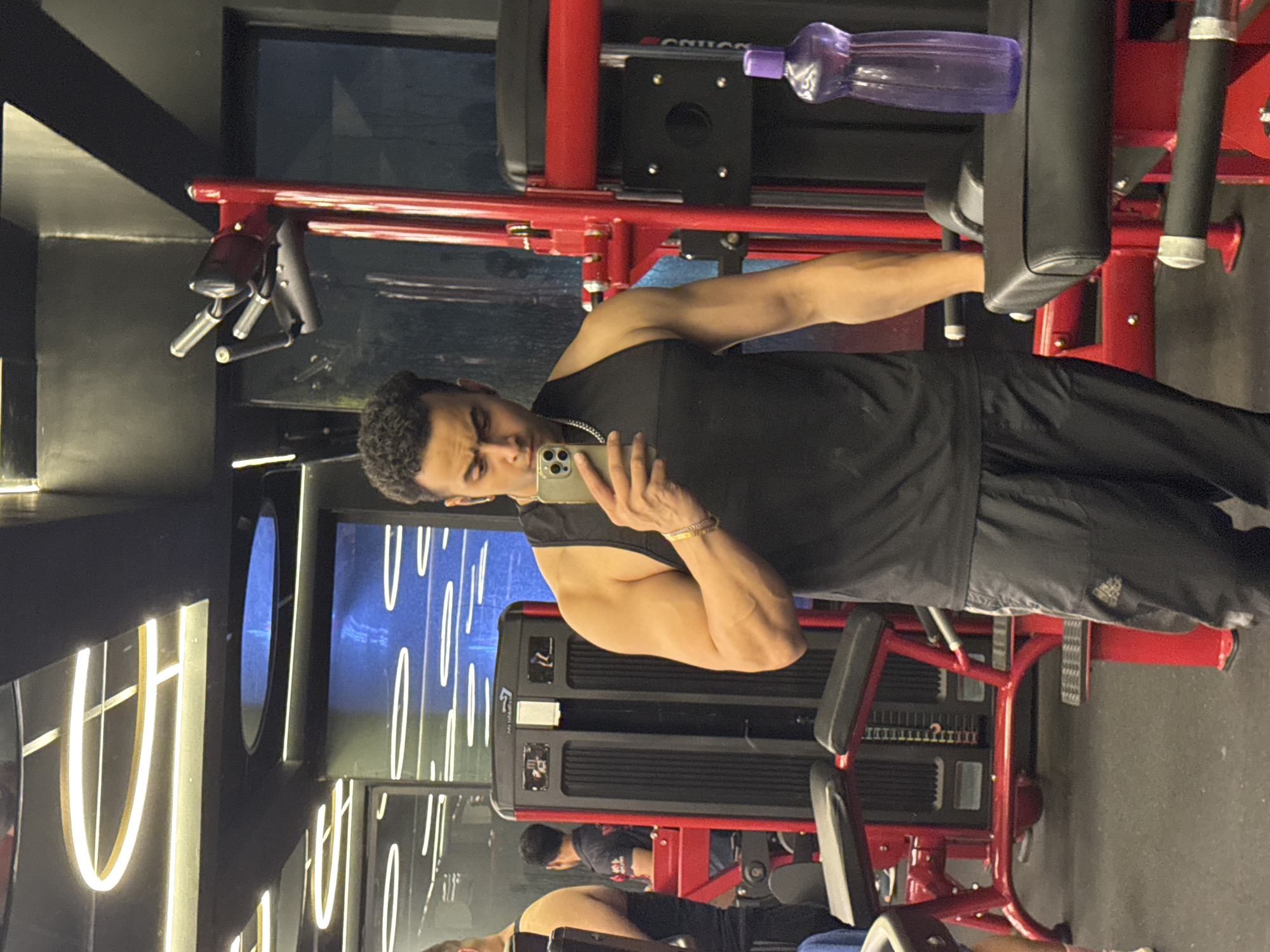 A man is taking a selfie in a gym. He is wearing a black tank top and black track pants. He has muscular arms and is wearing a silver chain around his neck and a bracelet on his left wrist. He is holding a silver iPhone in his right hand. The gym has red and black exercise equipment. In the background, there are other gym-goers and mirrors. The ceiling has modern lighting fixtures.