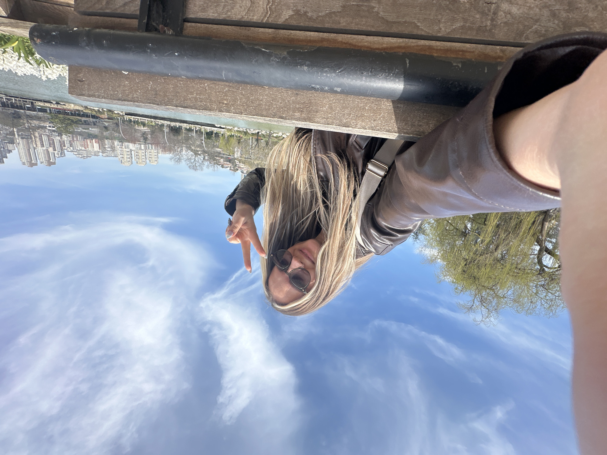 This is an outdoor wide-angle selfie captured on a clear day, depicting a location in Başakşehir, Turkey, consistent with Başakşehir Gölet Park. The primary subject is a female with long, blonde-streaked hair, positioned in the upper center of the frame. She wears dark sunglasses and a brown leather jacket, with a light-colored bag strap visible over her right shoulder. She exhibits a slight smile and holds her left hand up, forming a peace (V) sign. Her right hand, holding the camera, is partially visible in the extreme left foreground. In the foreground, a wooden bench or railing with a prominent dark, cylindrical metal bar extends horizontally. The background features a large body of water, likely an artificial lake or canal. Beyond the water, a dense urban landscape is visible on elevated ground, characterized by numerous modern, multi-story residential buildings. Scattered white flowers, indicative of spring bloom, are present along the water's edge in the lower right. A large tree with nascent light green leaves occupies the left side of the background. The sky is bright blue with scattered wispy white clouds.