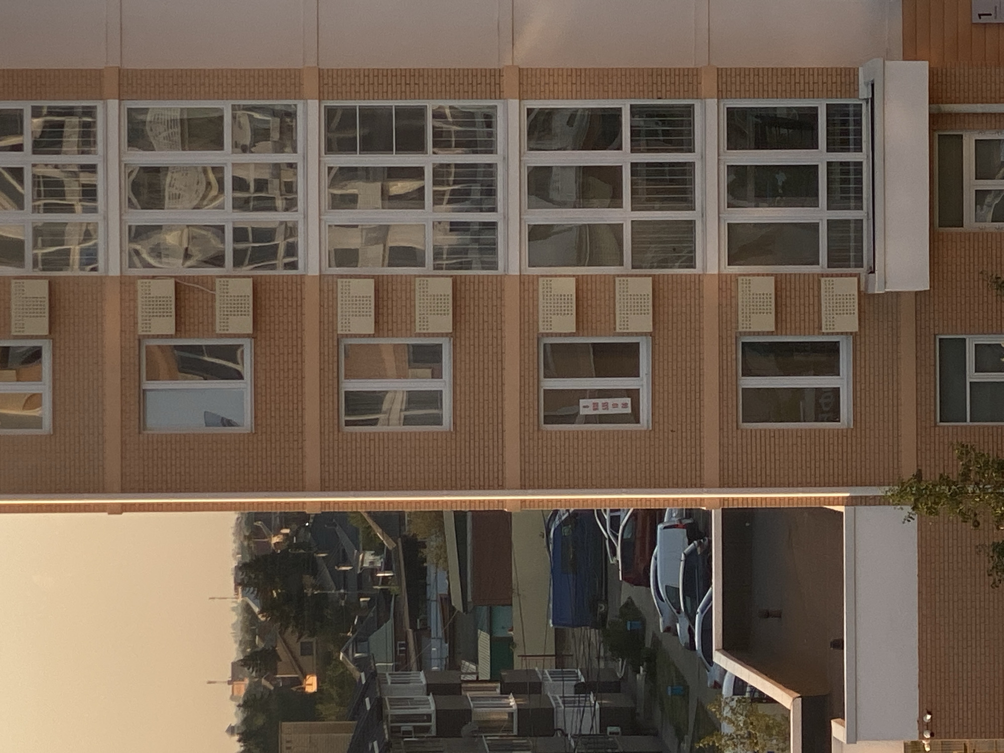 The image depicts a residential building in Agronom, Russia, featuring a brick facade and multiple windows. The building is tall, with several stories visible. The windows are large and appear to be modern, with white frames. Some windows have air conditioning units installed below them. The building is situated in a densely populated area, with other buildings and houses visible in the background, along with trees and a street with parked cars. The sky is hazy and appears to be either dawn or dusk. The overall scene suggests a typical urban or suburban environment.