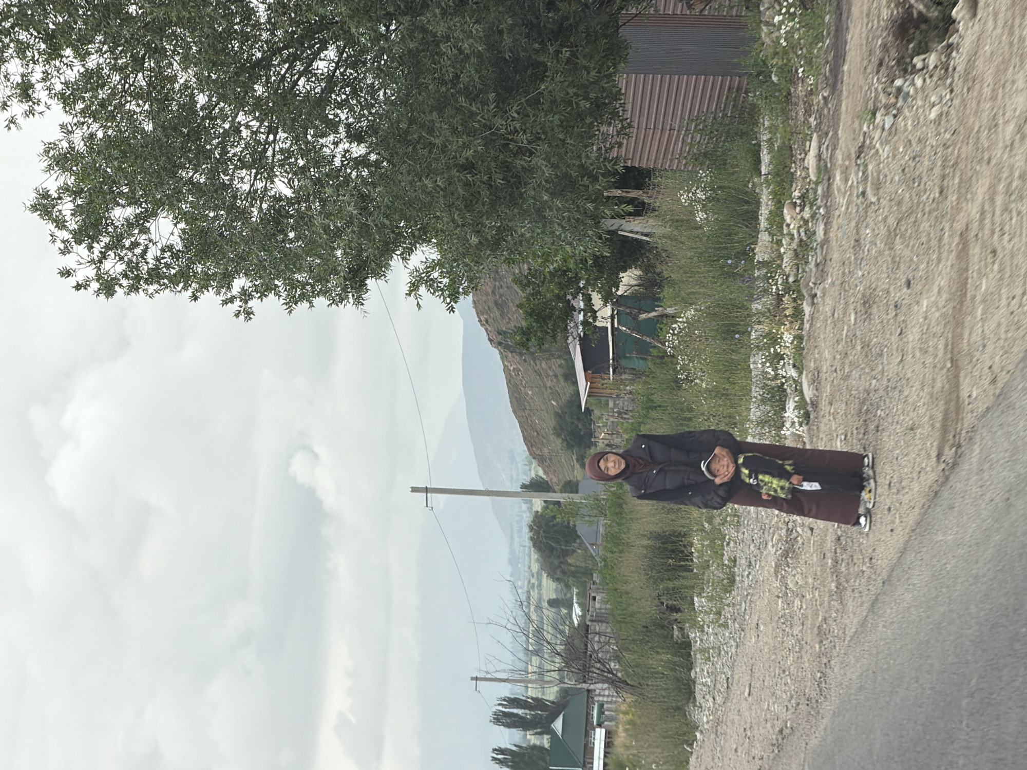 A medium-shot, eye-level, outdoor, vertical, natural-light photograph shows a woman and a child standing on a dirt road next to a gravel path. The woman, who is wearing a maroon hijab and a dark jacket, stands to the left of the frame, her hands touching the child's shoulders. The child, who is wearing a green, black, and yellow patterned jacket, stands in front of her, looking at the camera. The child's feet are on the gravel path. In the background, there are trees, houses, and mountains under a cloudy sky. The overall setting appears to be a rural or semi-rural area.