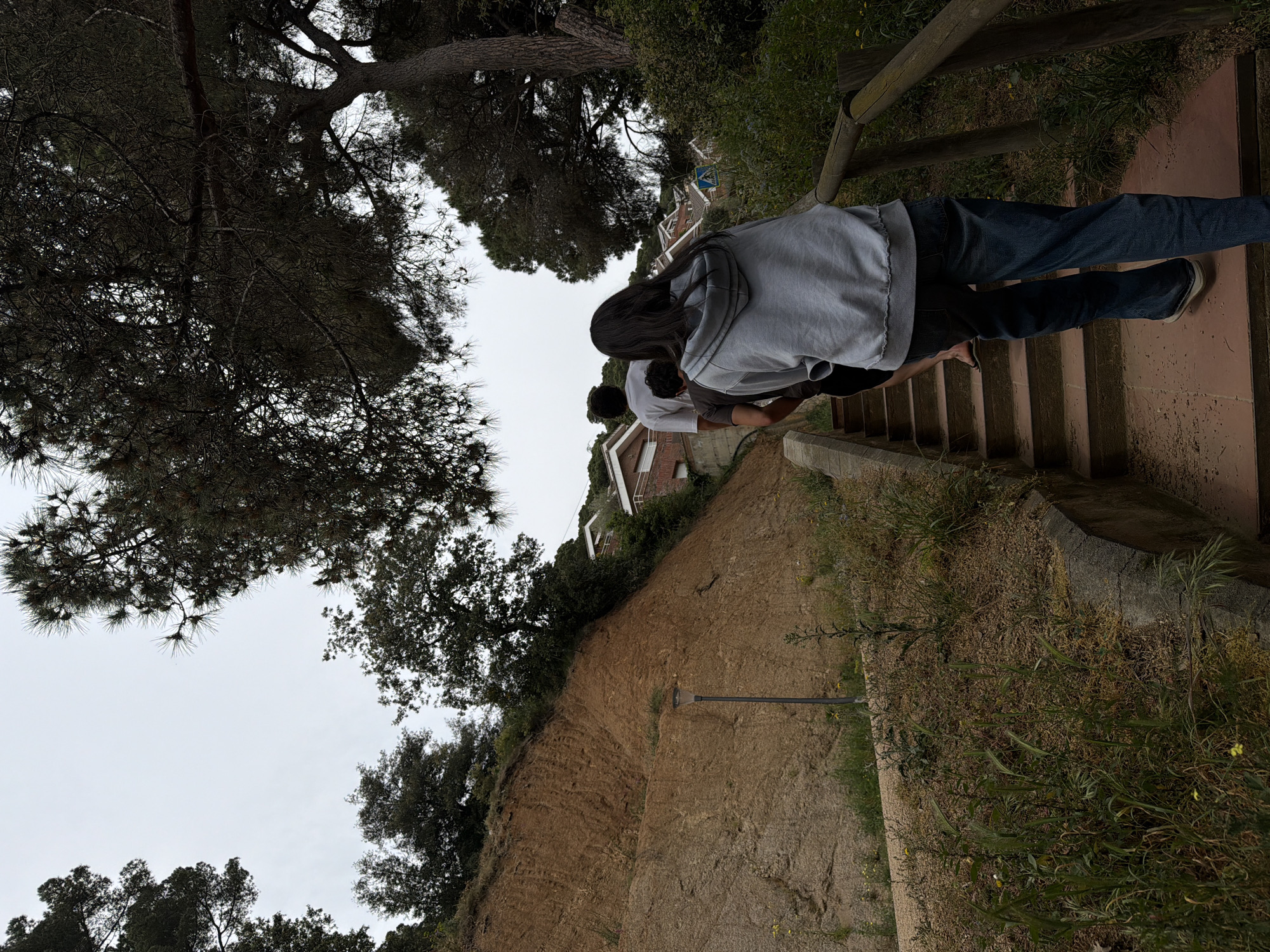 The image captures a scene in Sant Andreu De Llavaneres, Spain, featuring two individuals descending a set of outdoor stairs. The person in the foreground is wearing a grey hoodie and blue jeans. The second person, partially visible behind the first, is wearing a white t-shirt. The stairs are bordered on one side by a wooden railing. Beyond the stairs and railing, there's a steep, dirt embankment covered with vegetation. The background includes residential buildings partially obscured by trees and a cloudy sky. The stairs appear to be made of a reddish-brown material, possibly brick or tile. The surrounding environment includes trees, grass, and other plants.