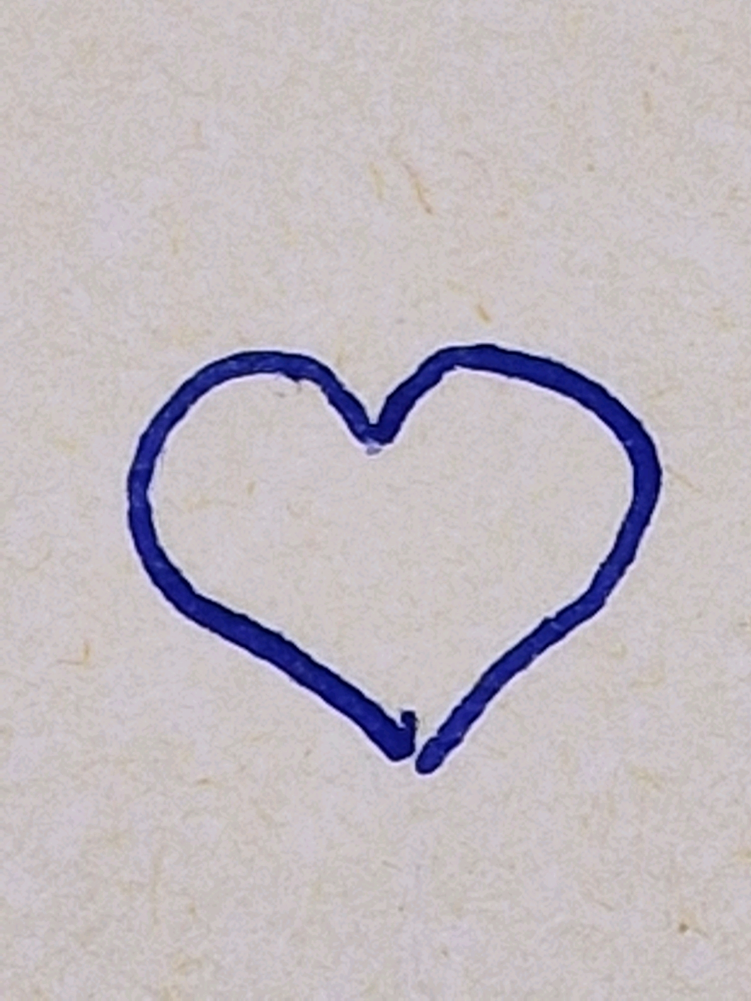 A blue, hand-drawn heart is centered on a textured, off-white background. The background appears to be paper, possibly handmade, with subtle variations in color and faint, darker fibers or speckles. The heart is drawn with a thick, uneven line, suggesting it was created with a marker or crayon. The drawing is not perfectly symmetrical, with the left side appearing slightly larger and more rounded than the right. There are no people, actions, or interactions depicted in the image, and the location is unknown.
