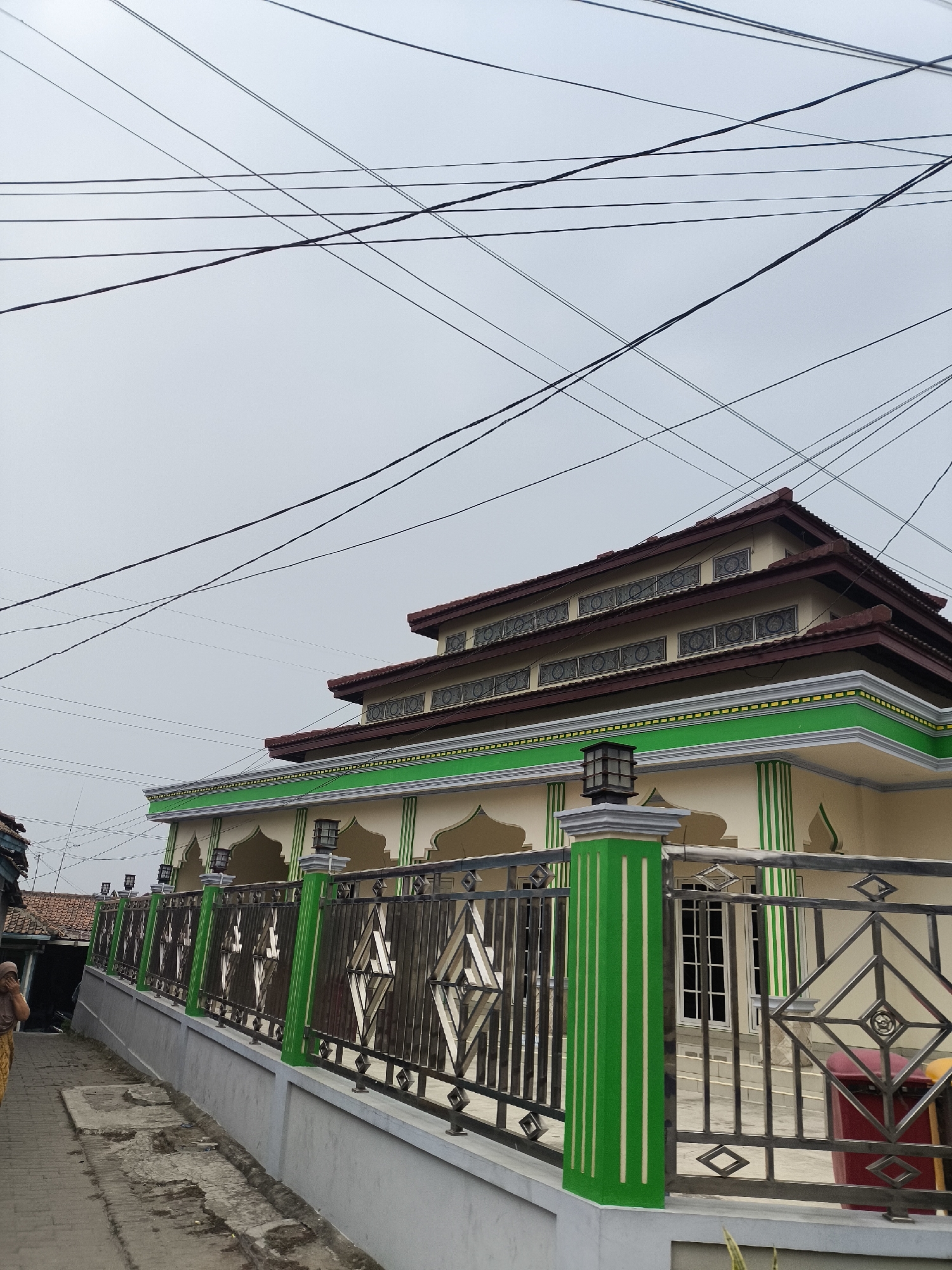 The image shows a mosque in Cilegon, Indonesia, under a cloudy sky. Overhead power lines crisscross the frame. The mosque has a multi-tiered, dark red tiled roof with decorative window frames. The facade is cream-colored with green accents, including a prominent green band running along the top of the wall and green stripes on pillars. A black metal fence with diamond-shaped insets and green-painted pillars surrounds the mosque. A person, partially visible on the left, is walking away from the camera on a paved sidewalk. The environment appears to be a residential area with other buildings visible in the background.