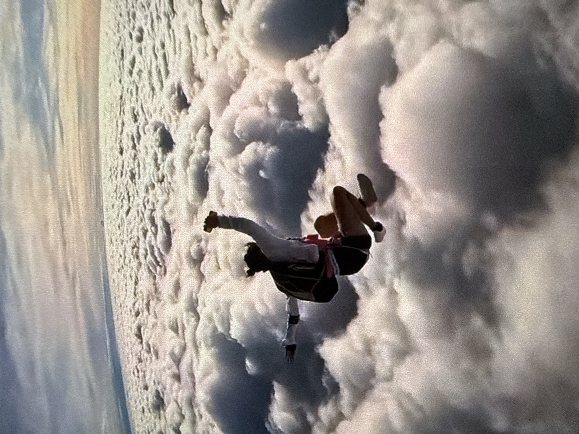 A person is freefalling in the sky amidst a dense cloud cover. They are wearing a white long-sleeved shirt, dark shorts, and shoes. A parachute harness is visible on their back. Their limbs are extended. The environment is dominated by the surrounding clouds with a blue sky in the distance. The location is unknown.