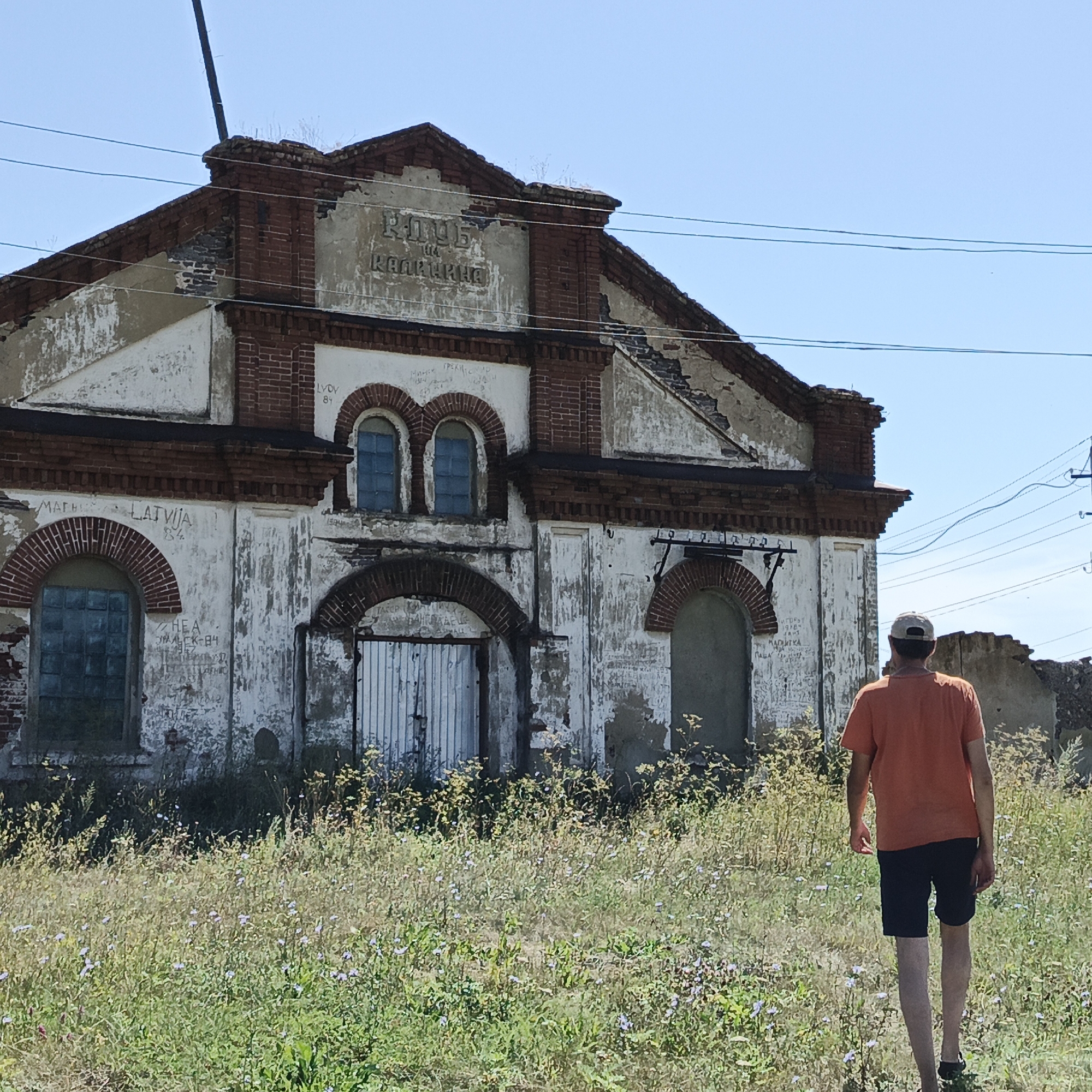 The image depicts a dilapidated brick and plaster building with peeling paint, located in the village of Tugustemir, Russia. The building shows signs of decay, with exposed brickwork and missing plaster. Above a central arched entrance, embossed lettering reads "КЛУБ КАЛИНИНА" which translates to "Kalinina Club". The building has multiple arched windows, some of which are boarded up or have glass panes. Graffiti is visible on the exterior walls.
In the foreground, a person wearing an orange t-shirt and dark shorts is walking away from the camera towards the building. They are wearing a baseball cap and sandals. The ground is covered in tall, dry grass and wildflowers. A utility pole with overhead wires is visible in the background to the right of the building. The sky is clear and blue. The scene suggests an abandoned or disused structure in a rural setting.