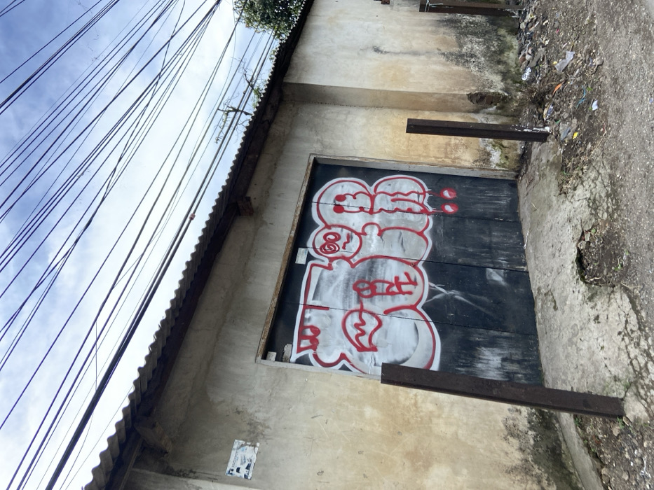 The image shows a concrete building with a large wooden door covered in graffiti. The door is a dark grey, with large white letters sprayed on it, as well as some red details.  The building is located on a dirt road, and is surrounded by an unkempt overgrown area with debris and trash scattered around. The building has a concrete roof with a corrugated metal awning. There are numerous power lines overhead, running parallel to each other. The building appears to be located in a small town or village. The building is in Soreang, Indonesia at the coordinates -7.033 latitude and 107.503 longitude.  No people are visible.