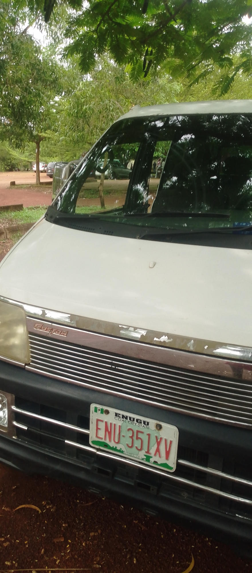 The image shows the front of a white van parked outdoors. The van has a chrome grille and a license plate. The license plate is white with green lettering and says "ENU-351XV" and "ENUGU COAL CITY STATE". The van is parked on a dirt surface, and there are trees and other vehicles visible in the background. The leaves of a tree are hanging over the top of the van, suggesting it is daytime and the weather is likely pleasant. The scene appears to be a parking area or roadside in a rural or semi-rural location in Nigeria, given the license plate information. There are no people prominently featured in the image, though faint figures can be seen reflected in the van's windows. The van appears to be slightly aged, with some peeling paint on the chrome trim above the grille. The overall impression is a straightforward shot of a parked vehicle in an outdoor setting.