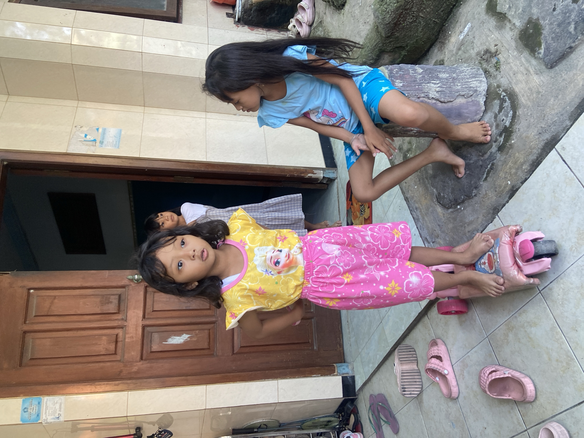 The image depicts an outdoor scene at a residence in Gelam, Indonesia. In the foreground, a young girl with dark hair and a yellow t-shirt adorned with an image of Elsa from Frozen, and pink patterned shorts, stands on a pink toy scooter. Her hands are on her hips. To her right, another young girl with long dark hair, wearing a blue t-shirt with a unicorn graphic and blue shorts with white stars, sits on a rough, cylindrical stone block. She is looking to her right. In the background, partially visible through an open wooden door, another child in a white top and a patterned skirt is standing. The ground is tiled and appears wet in places. Several pairs of pink flip-flops are scattered on the floor around the girls. The walls are tiled with a light-colored square pattern. To the left of the door, a badminton racket is leaning against the wall.