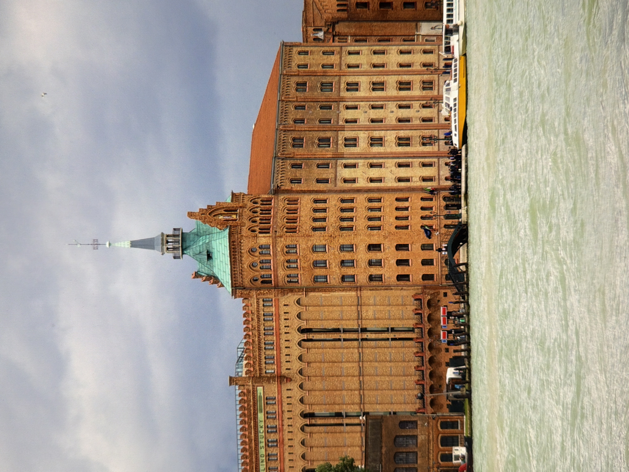 A wide-angle, slightly hazy, medium-distance photograph captures a large brick building with a green-tiled roof and a tall, slender spire. The building features numerous windows and decorative architectural elements, suggesting it is an older, historic structure. In front of the building, a body of water, likely a canal or river, is visible, with several boats docked or in motion. The sky above is overcast with dark clouds, and a small bird is visible in the upper right quadrant. The scene appears to be an urban waterfront with notable architecture. The building bears a sign that reads "LINO STUCKY." This building is the former Lino Stucky grain mill, a prominent landmark in Venice, Italy, not Kyiv, Ukraine.