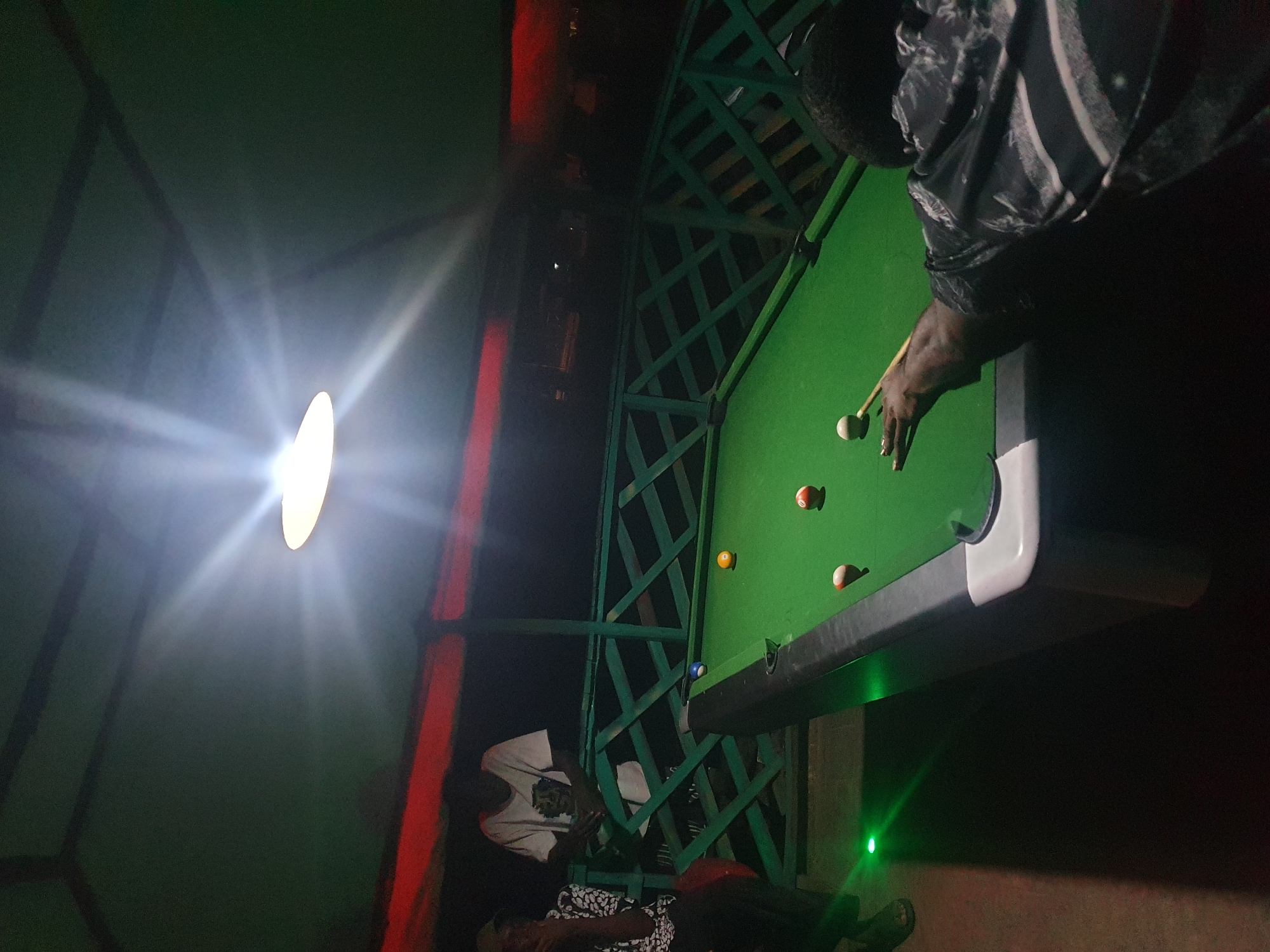The image depicts an outdoor pool hall at night, likely in Lokoja, Nigeria, illuminated by a bright overhead light. A pool table with green felt occupies the foreground, with several colored balls scattered across it. A person is actively playing pool, leaning over the table with a cue stick in hand. In the background, two other individuals are visible, one standing and observing the game, and another seated. The setting appears to be a casual, possibly recreational, outdoor establishment with a green railing in the middle ground and a dark, indistinct background suggesting the surrounding environment. Green lights are also visible on the ground, adding to the ambiance.