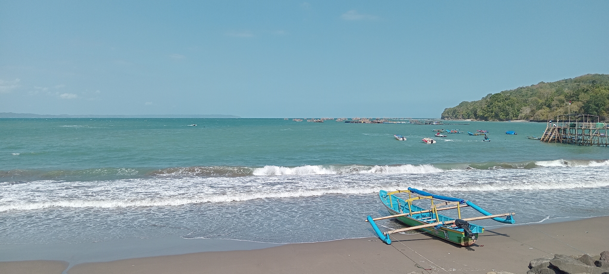 The image depicts a coastal scene at Desa Pagandaran, Indonesia. The foreground features a sandy beach with a small, brightly colored blue and green fishing boat resting on the shore. The boat has outriggers for stability and a small outboard motor attached to its stern. Gentle waves are breaking on the dark sand beach, and the water transitions to a clear turquoise hue further out.
In the mid-ground, the sea is dotted with numerous small fishing boats and floating structures, likely fish farms or aquaculture pens, stretching across the horizon. A few faster boats, possibly speedboats, are also visible. To the right, a wooden stilted structure, possibly a pier or viewing platform, extends into the water, with a few people on it.
The background consists of a lush, tree-covered hillside that slopes down to the sea, creating a natural boundary for the bay. The sky is a clear, pale blue with a few wispy white clouds. The overall impression is a calm, sunny day at a coastal fishing village.