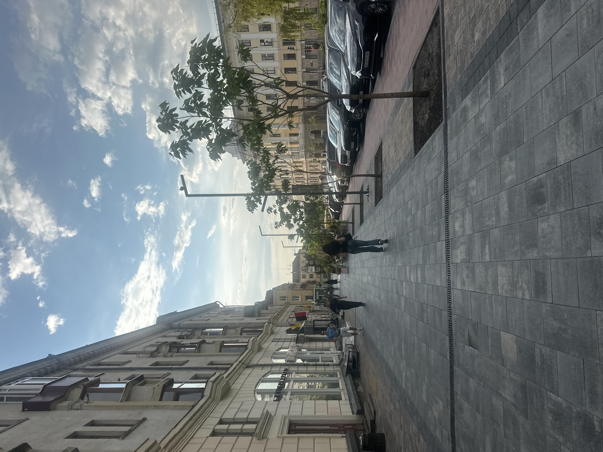 The image depicts a street scene in Ternopil, Ukraine. On the left side of the image, a light-colored building with multiple stories and numerous windows runs along the street. The ground level of the building features storefronts with large windows and doors. A sidewalk, paved with dark grey rectangular tiles, extends along the building. Several people are walking on the sidewalk. To the right of the sidewalk, a row of parked cars is visible, mostly dark-colored sedans. A line of young trees with green foliage is planted along the edge of the sidewalk, separating it from the parked cars. In the background, more buildings and trees are visible. The sky is blue with scattered white clouds. The overall impression is that of a well-maintained urban street on a pleasant day.