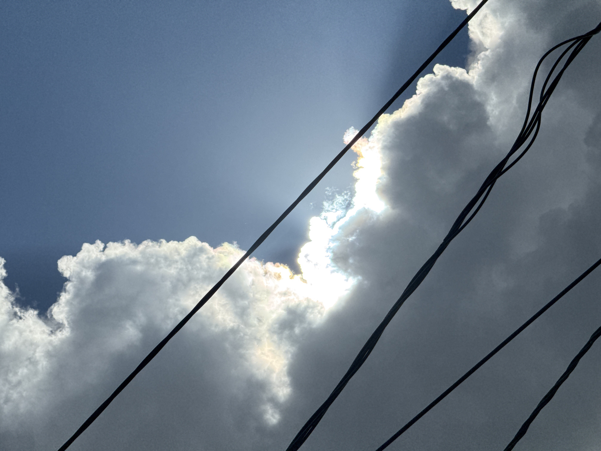 The media file captures a dramatic sky with the sun peeking through billowing clouds. The sun's rays create a radiant halo effect, illuminating the edges of the clouds with a bright, almost iridescent glow. Several dark, diagonal lines of what appear to be electrical wires crisscross the frame, adding a graphic element and a sense of depth to the composition. The sky itself is a mix of deep blue and overcast gray, suggesting dynamic weather conditions.
The overall mood is one of awe and perhaps a touch of foreboding, due to the contrast between the bright sun and the dark, imposing wires against a darkening sky. The scene is likely set outdoors, looking upwards. The time of day is difficult to pinpoint precisely, but the quality of light suggests it could be late afternoon or early evening, when clouds can often catch the sun in such striking ways. There is no visible text in the image. The setting implies an urban or semi-urban environment, given the presence of electrical wires. The weather appears to be changing, with the possibility of impending rain or a passing storm, yet with moments of intense sunlight breaking through.