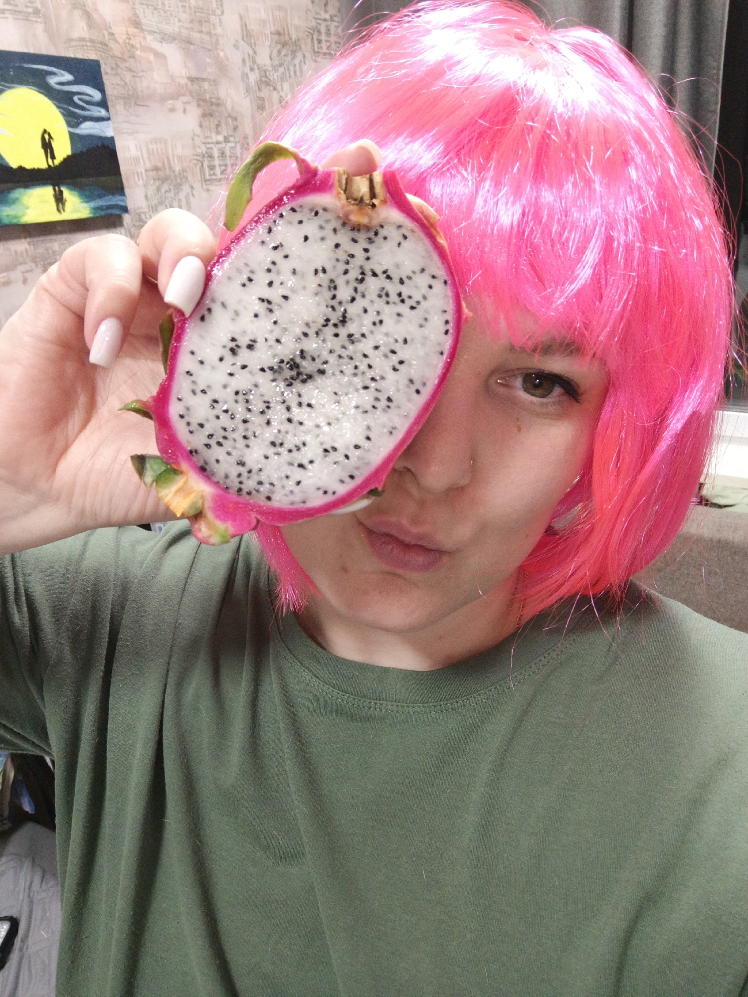 A person with fair skin and brown hair, wearing a short, bright pink wig, holds a dragon fruit half in front of their face. The dragon fruit is pink on the outside, with green leaves, and white on the inside with black seeds. The person is wearing a green t-shirt and has a nose piercing in their left nostril. They are making a slight pouting face. Their nails are painted light pink. In the background, there is a painting of a yellow moon with two figures standing on land reflected in water. The location is Марковский, Russia.