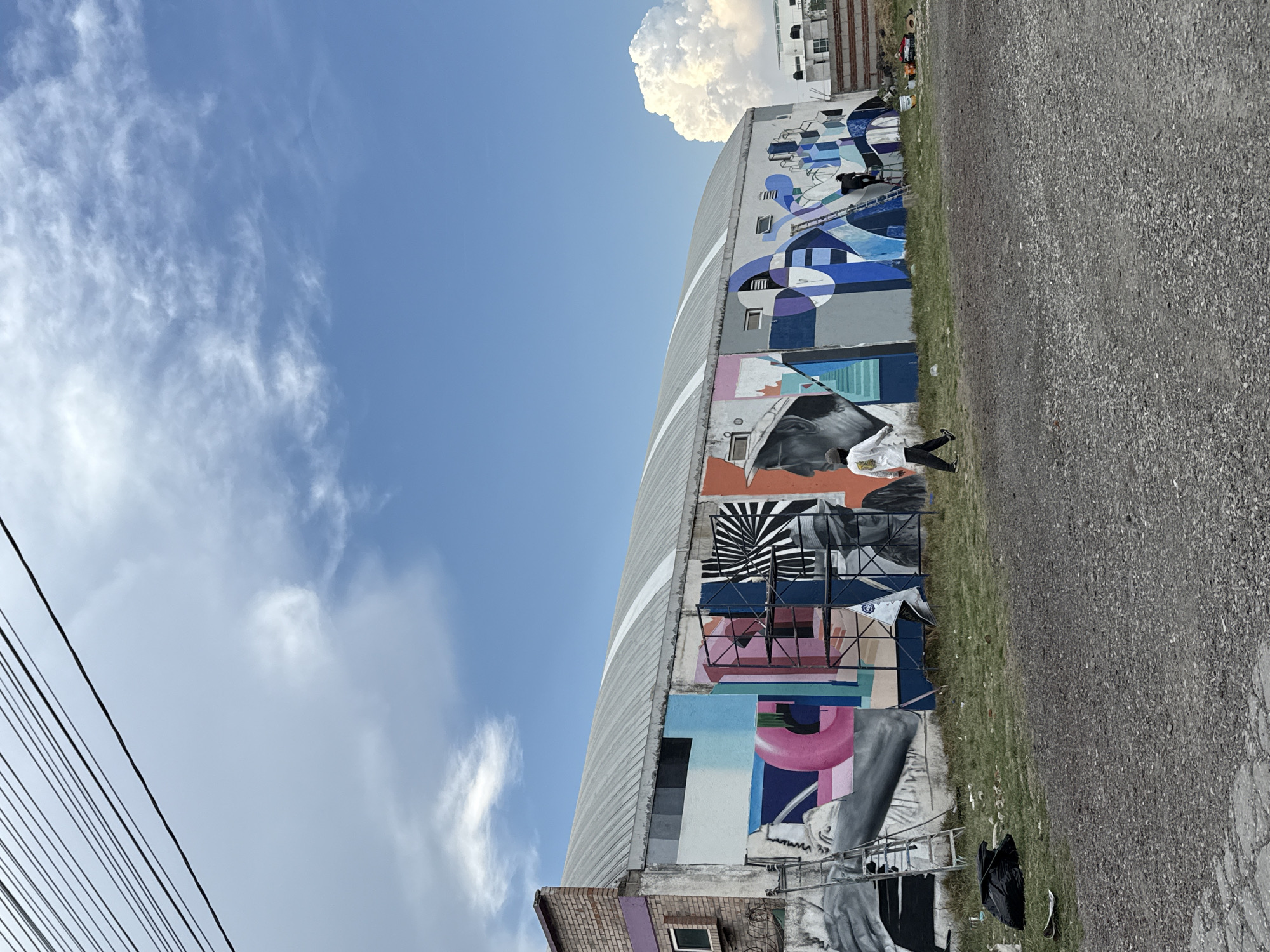 The image captures a building in San Andrés Cholula, Mexico, featuring a large-scale mural spanning multiple sections. The mural includes a variety of styles and subjects, ranging from abstract geometric designs and monochromatic portraits to scenes depicting people and objects. A person is walking near the base of the building, adjacent to a grassy area that separates the mural from a paved road. Utility lines are visible in the lower left corner of the frame. The sky overhead shows a mix of blue with scattered clouds.