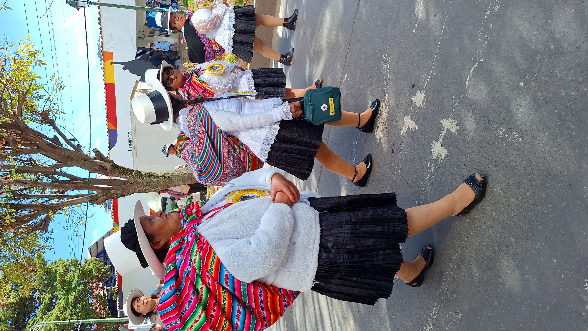 Four women wearing traditional Andean attire, including wide-brimmed white hats and colorful woven shawls, walk down a paved street in Wanchaq, Peru. The woman in the foreground on the left has a white fuzzy jacket and a black pleated skirt. The woman next to her in the center is wearing a white lace blouse and a black pleated skirt, carrying a small green medical bag. To her right, another woman is dressed similarly, and the fourth woman on the far right wears a black hat with white trim. They are walking in the same direction, suggesting a procession or parade. The background features buildings with a mural depicting a figure with an outstretched arm and the word "UNIDOS". A tree with green foliage and a bright blue sky with scattered clouds complete the scene.