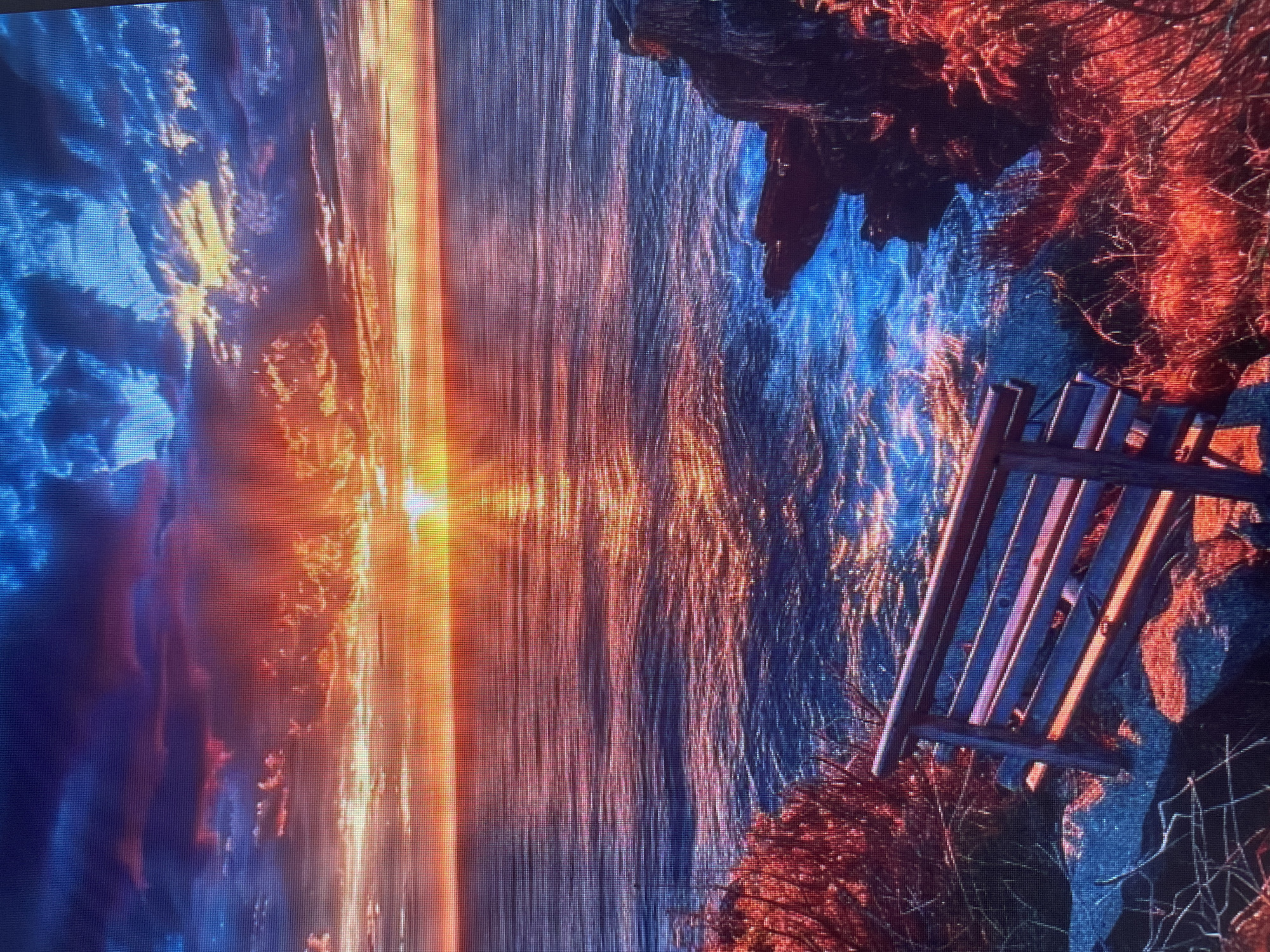 The image depicts a dramatic sunset over a body of water, with a bench in the foreground overlooking the scene. The sky is a vibrant mix of oranges, reds, blues, and purples, with clouds catching the light of the setting sun. The sun itself is low on the horizon, casting a brilliant golden pathway across the rippling water. To the right, rocky cliffs and reddish-brown vegetation frame the view. In the foreground, a wooden bench with slatted seats and backrests sits on a rocky outcrop amidst sparse, dry-looking grass. The overall impression is one of serene, natural beauty, amplified by the intense colors of the sunset. The location is not specified as Sinyavino, Russia; this is a general scenic landscape.