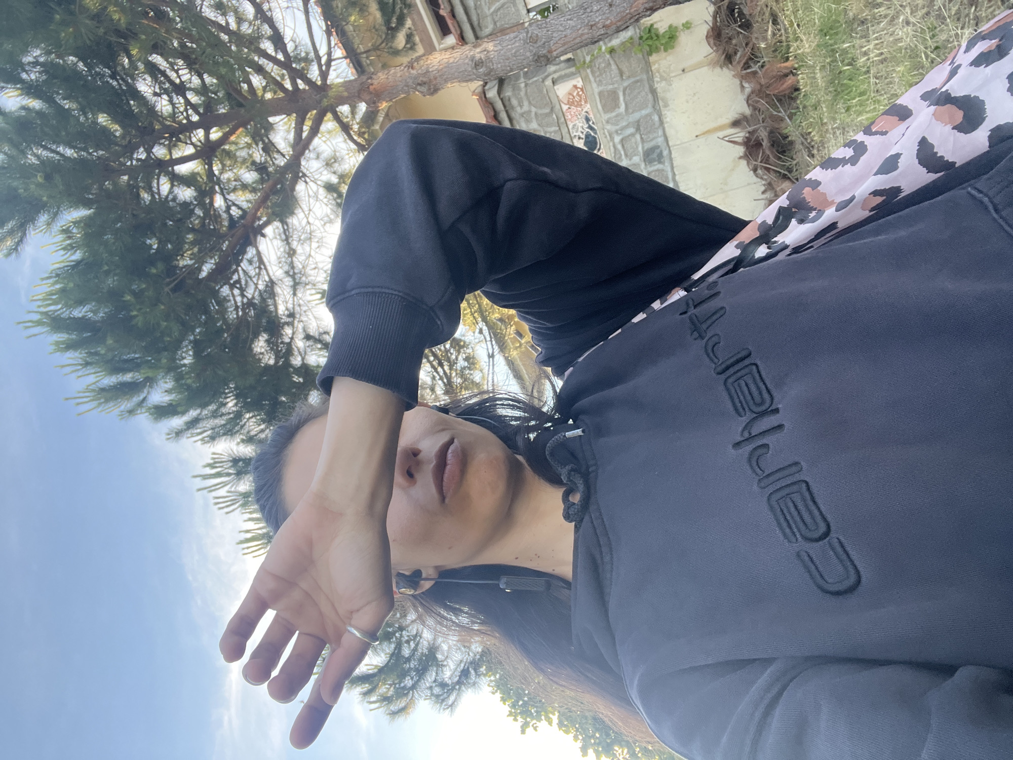 A selfie taken outdoors on a sunny day shows a person with long, dark hair covering their left eye with their right hand. They are wearing a dark grey hooded sweatshirt with the word "carhartt" embroidered in black on the front. A leopard print scarf or fabric is visible peeking out from under the sweatshirt on the right side. The person is also wearing earbuds. The background features a tall pine tree, a stone building with a red tile roof, and dry vegetation on the ground. The sky above is blue with some light clouds. The scene is consistent with an outdoor setting in Sinemorets, Bulgaria, known for its natural beauty and coastal landscapes.