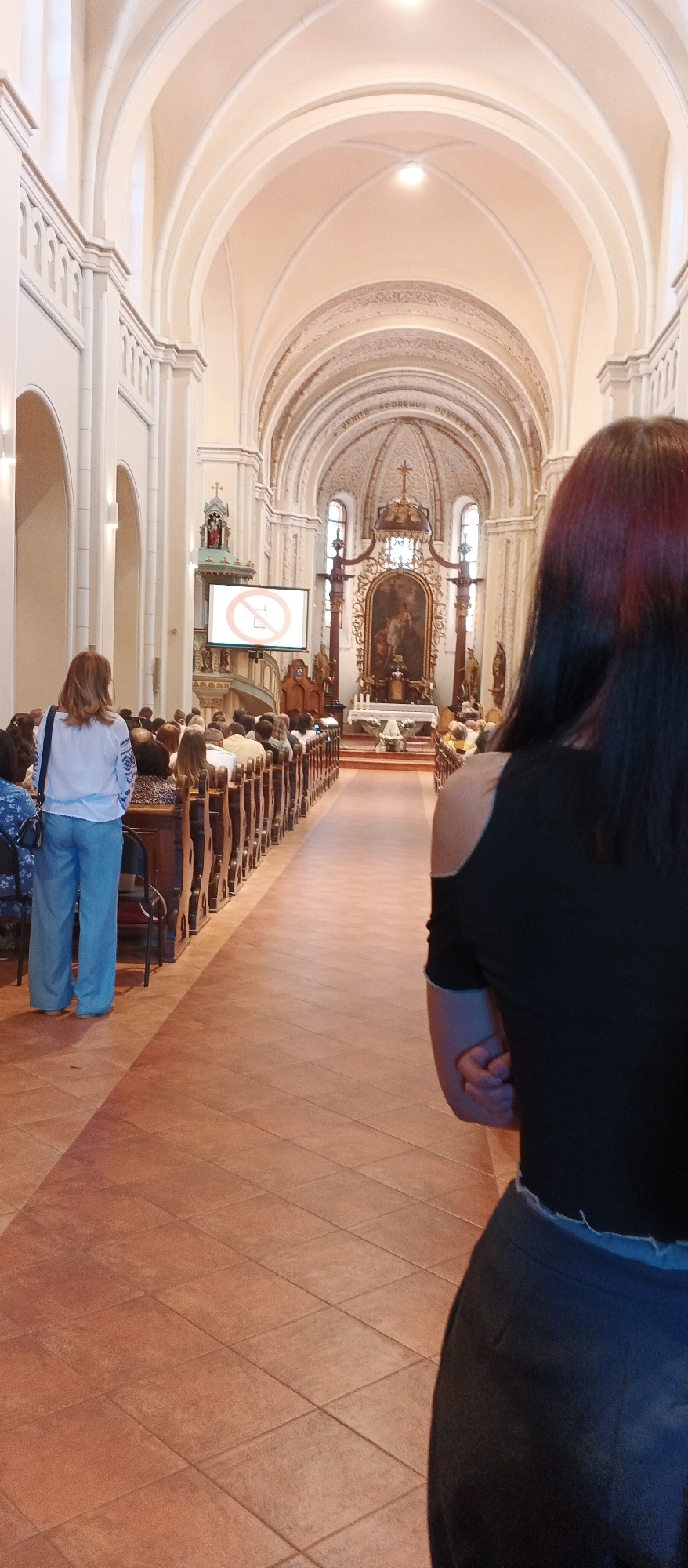 The image depicts the interior of a church, likely St. Martin's Cathedral in Mukachevo, Ukraine, given its architectural style and religious setting. The perspective is from the back of the church, looking towards the altar.
In the foreground, on the right, a person with dark hair is seen from the back, wearing a black top with exposed shoulders and dark pants. To the left, a woman with reddish-brown hair is standing, facing away from the camera and towards the altar. She is wearing a light-colored, embroidered blouse and wide-legged blue trousers.
The church interior features rows of wooden pews filled with a congregation. A screen is visible on the left side of the nave displaying a red circle with a diagonal line through a mobile phone symbol, indicating a no-phone policy. The architecture is grand, with high arched ceilings, ornate decorative elements, and stained glass windows. The floor is tiled in a reddish-brown color. At the front of the church, the altar area is adorned with religious artwork, candles, and decorations, illuminated by overhead lights. The overall atmosphere suggests a religious service or gathering is in progress.