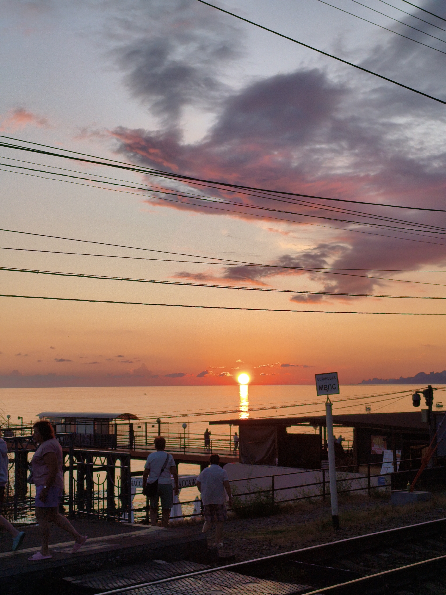This is a vertical, eye-level, outdoor, daytime photograph taken at sunset in Sochi, Russia. The scene is dominated by a vibrant orange and pink sunset over the Black Sea, with the sun a bright orb on the horizon. Several people are visible, walking on a pier and along a walkway beside train tracks. The pier has a covered waiting area and fencing. A sign with Cyrillic lettering reads "ОСТАНОВКА МВпс". Overhead, numerous dark power lines crisscross the sky. The overall atmosphere is serene and picturesque, with the warm hues of the sunset casting a soft glow over the environment.