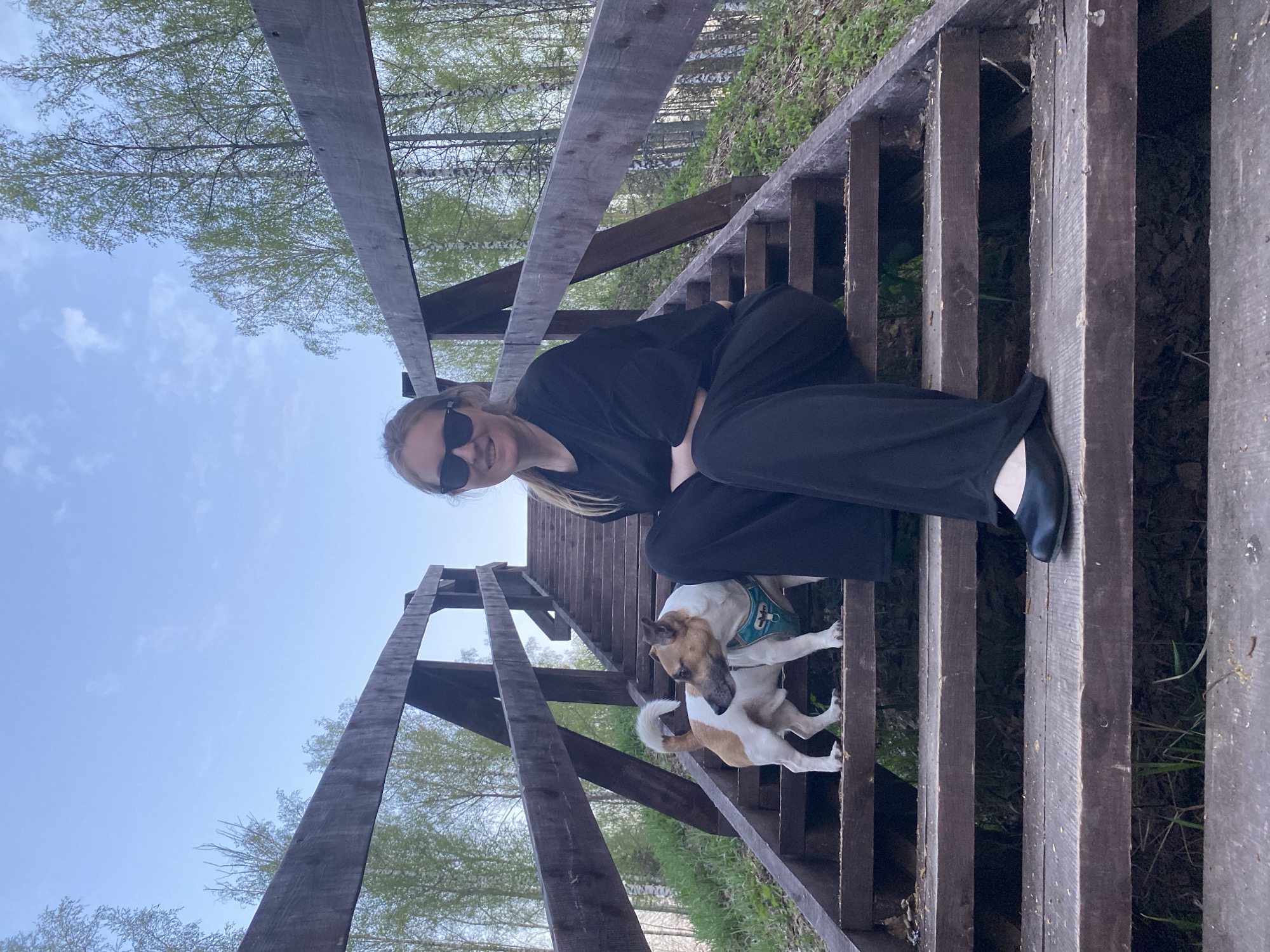 A woman and two dogs are on a wooden staircase, outdoors, in Noviy, Russia. The woman is fair-skinned, wearing sunglasses and a black outfit, and is sitting on a step of the staircase. Below her are two tan and white dogs. The staircase is made of weathered wood and leads upwards. Trees are visible in the background, with a blue sky overhead.