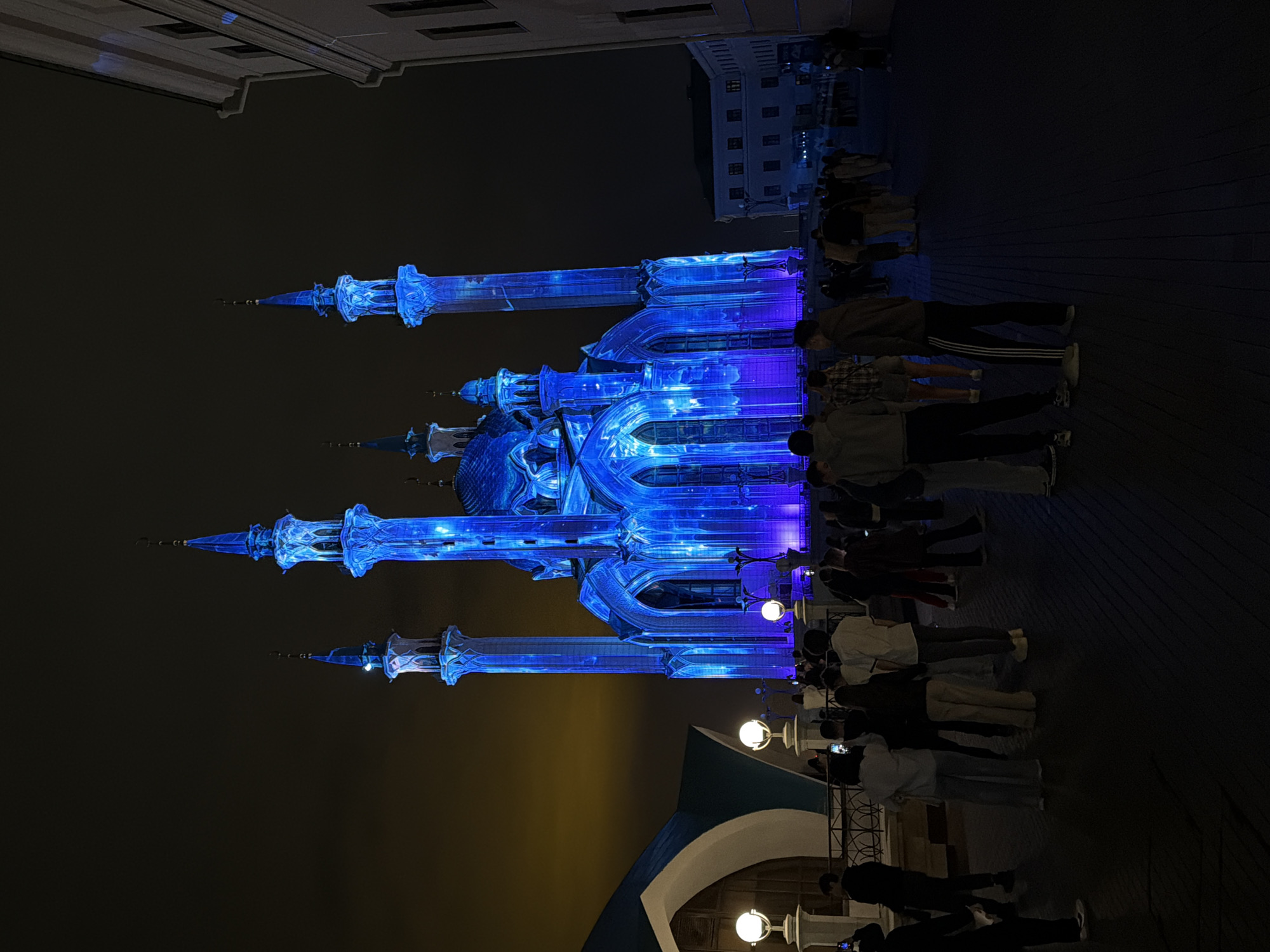 At night, the illuminated Qolşärif Mosque in Kazan, Russia, stands brightly, its minarets and dome bathed in a vibrant blue light. A crowd of people gathers in front of the mosque, many holding up their phones to take pictures of the spectacle. Streetlights dot the foreground, adding points of light to the scene. Buildings are visible in the background, silhouetted against the dark sky.