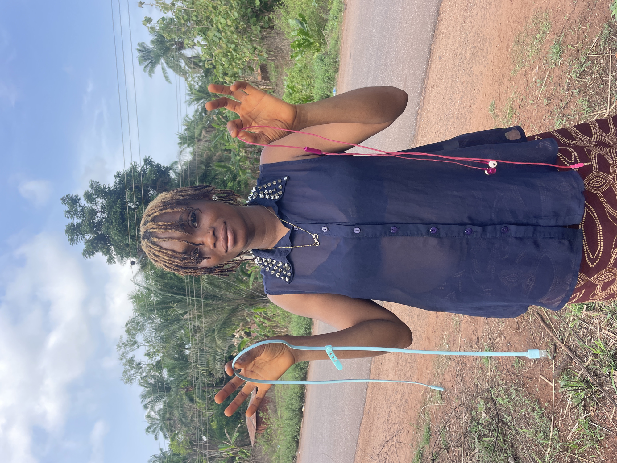 A woman stands on the side of a paved road in Ogbe, Nigeria. She wears a navy blue, semi-sheer button-down shirt with silver studs on the shoulders, and a patterned skirt. She holds a light blue USB cord in one hand and pink wired earphones in the other. The background shows a roadside with vegetation and trees, with a cloudy sky above. Power lines are visible overhead.